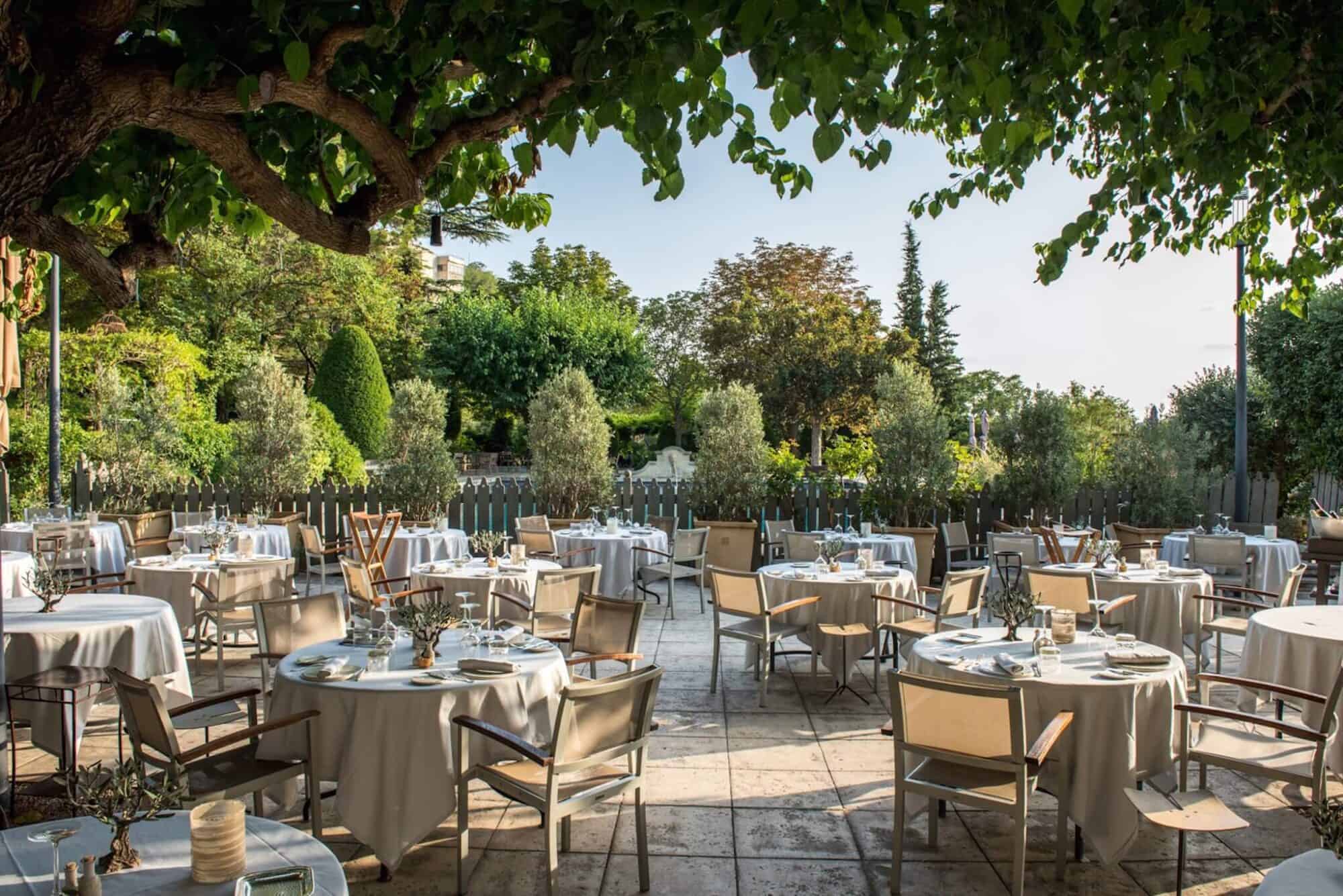 Tables and chairs are set out on a wide, sunny porch under leafy green trees at Baumanière in Arles. 