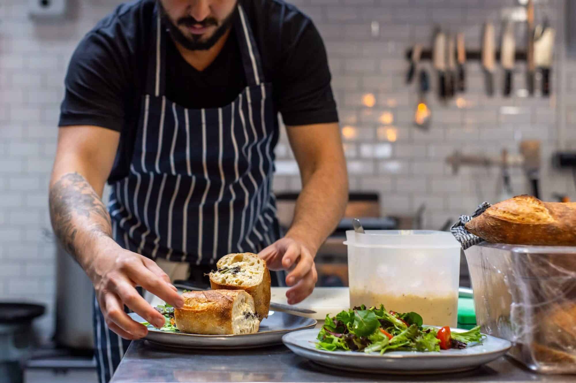 A chef wearing a striped apron in a professional kitchen plates a sandwich, ready to be served to customers in a restaurant. There is a plate of salad next to the sandwich plate, and knives can be seen in the distance on the wall behind the chef.