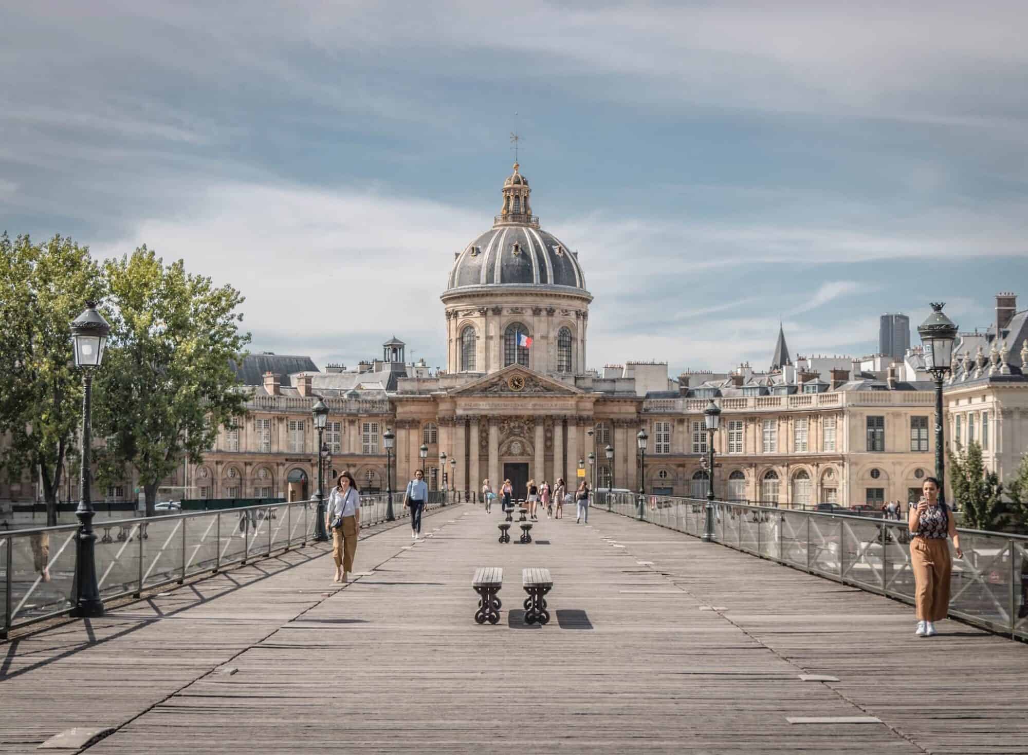 A view of the Institut de France from the Pont des Arts bridge in Paris, where people are seen walking on either side of the bridge.