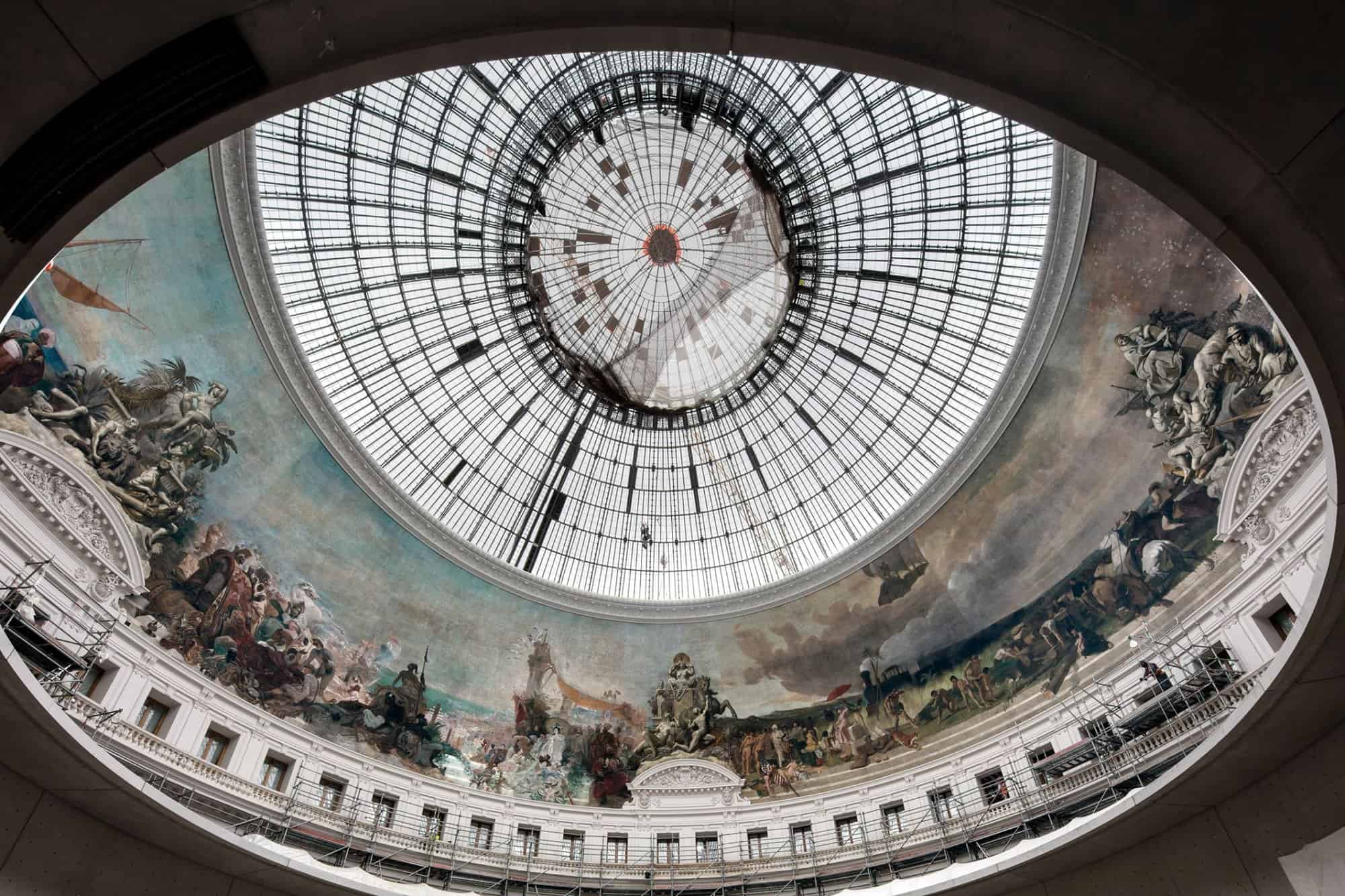 A large dome ceiling made of glass. There is a large mural below the glass ceiling. There are white walls under the mural with scaffolding, and there is a net on the glass ceiling for construction work