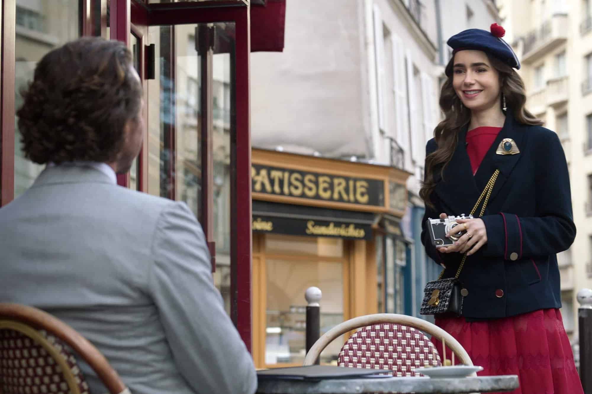 A woman with brown hair wearing a blue beret, blue jacket, and a red dress standing in front of a man with his back to the camera sitting outside at a café. A pastry shop with "patisserie" written on it can be seen in the background. The woman is Emily from "Emily in Paris."