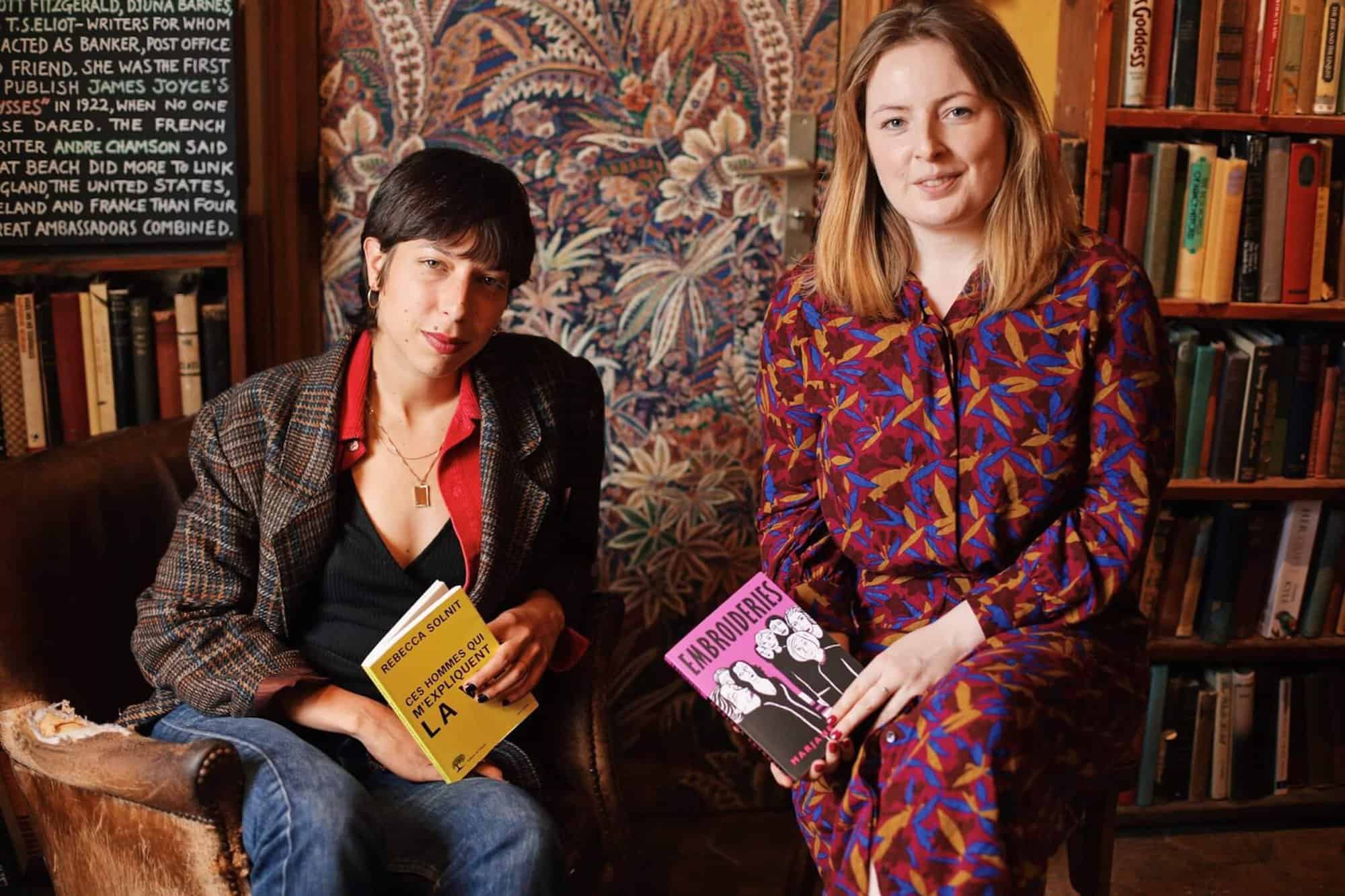 Hosts of the Feminist book club, Camille Lou to the left and Lou Binns to the right, sitting side by side with books in their hands. Camille dressed in a blazer, tshirt and jeans, Lou wearing a long, printed dress, and the two smile at the camera.
