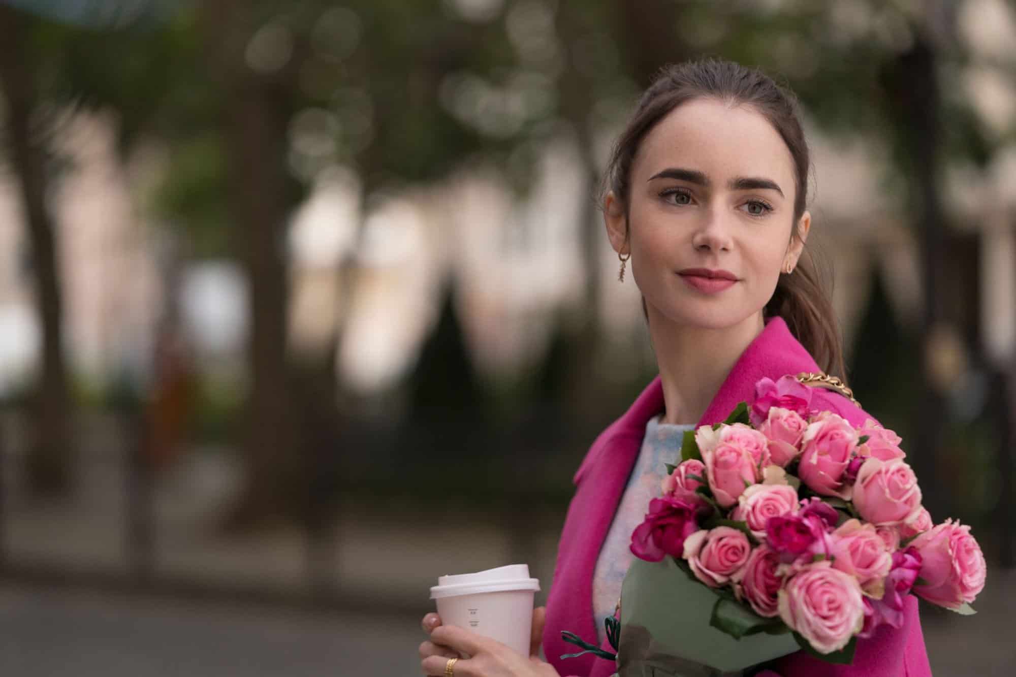A woman with her hair pulled back wearing a bright pink blazer carrying a bouquet of pink flowers. She is also carrying a cup of coffee to-go and smiling into the distance.