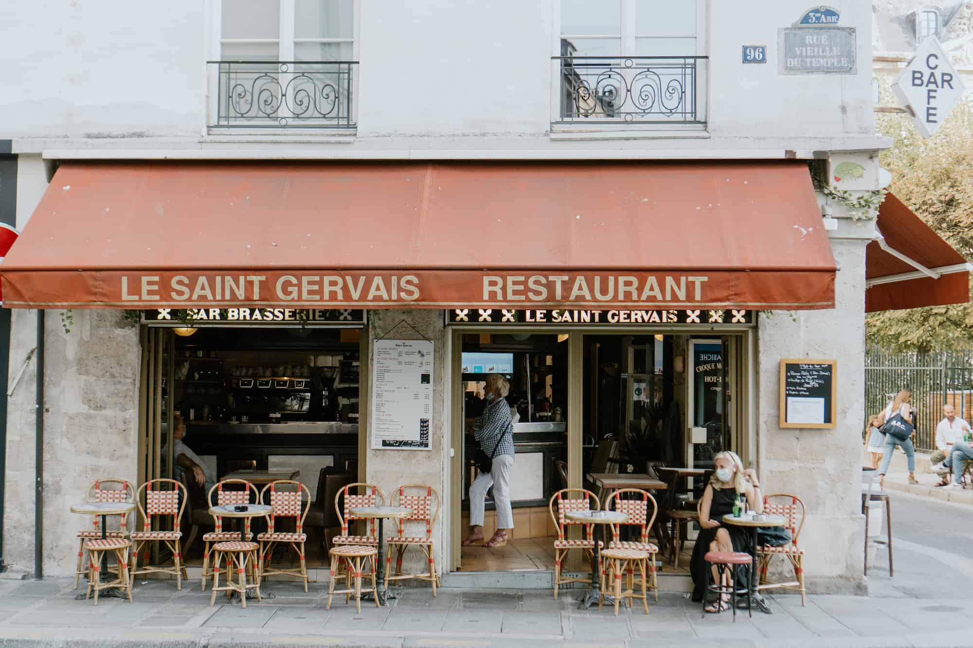 The exterior of a café in Paris. The awning is orange and reads "Le Saint Gervais Restaurant." There is a woman wearing a mask seated at one of the tables in front of the café. There is another woman standing at the bar who is visible inside the café.