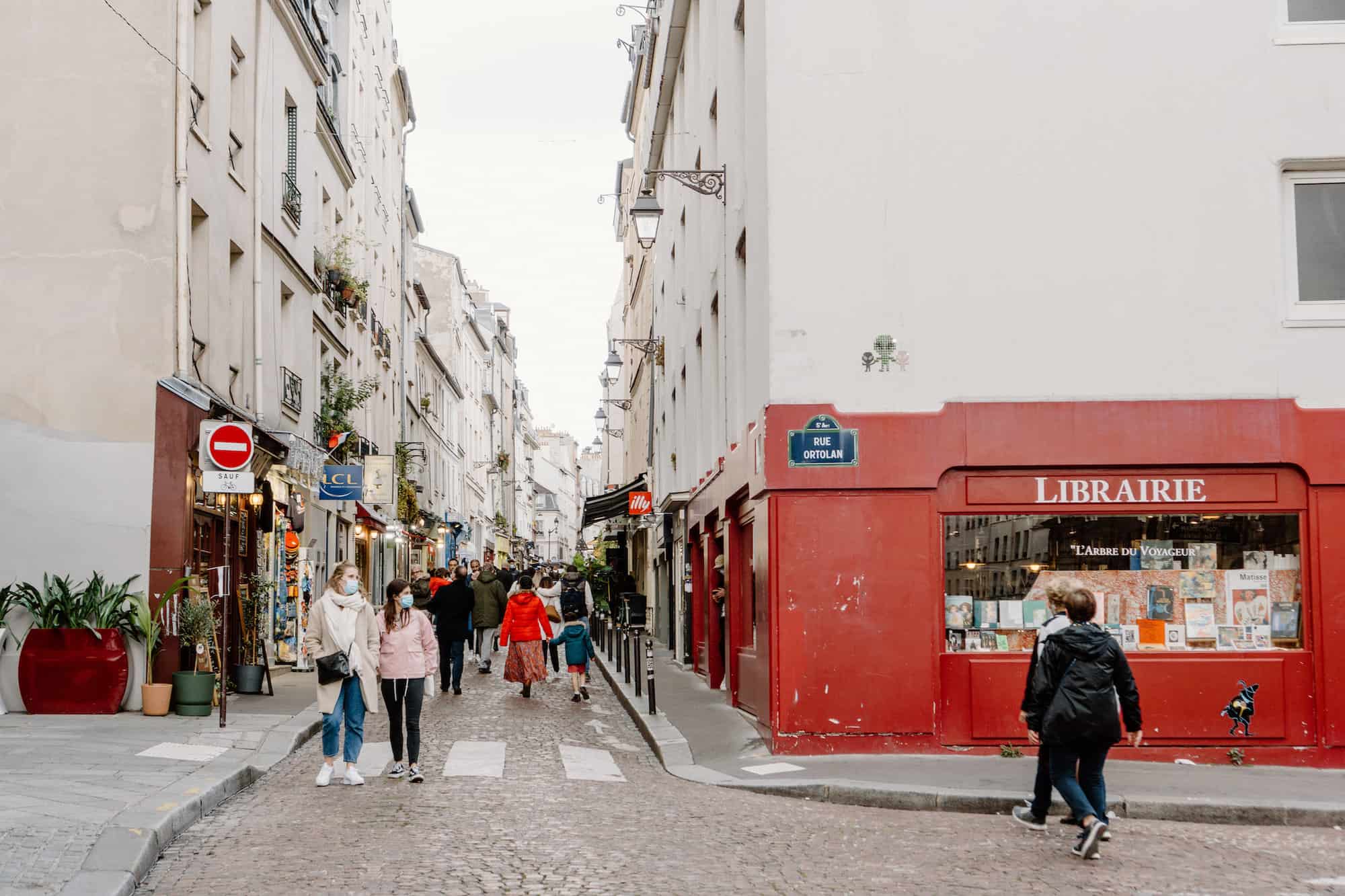 A street in Paris. There is a red bookstore with the word "Librarie" written in white on it to the right. There are several people walking in the street, all who are visible are wearing face masks. There is a do not enter street sign visible to the left. The surrounding buildings are white.