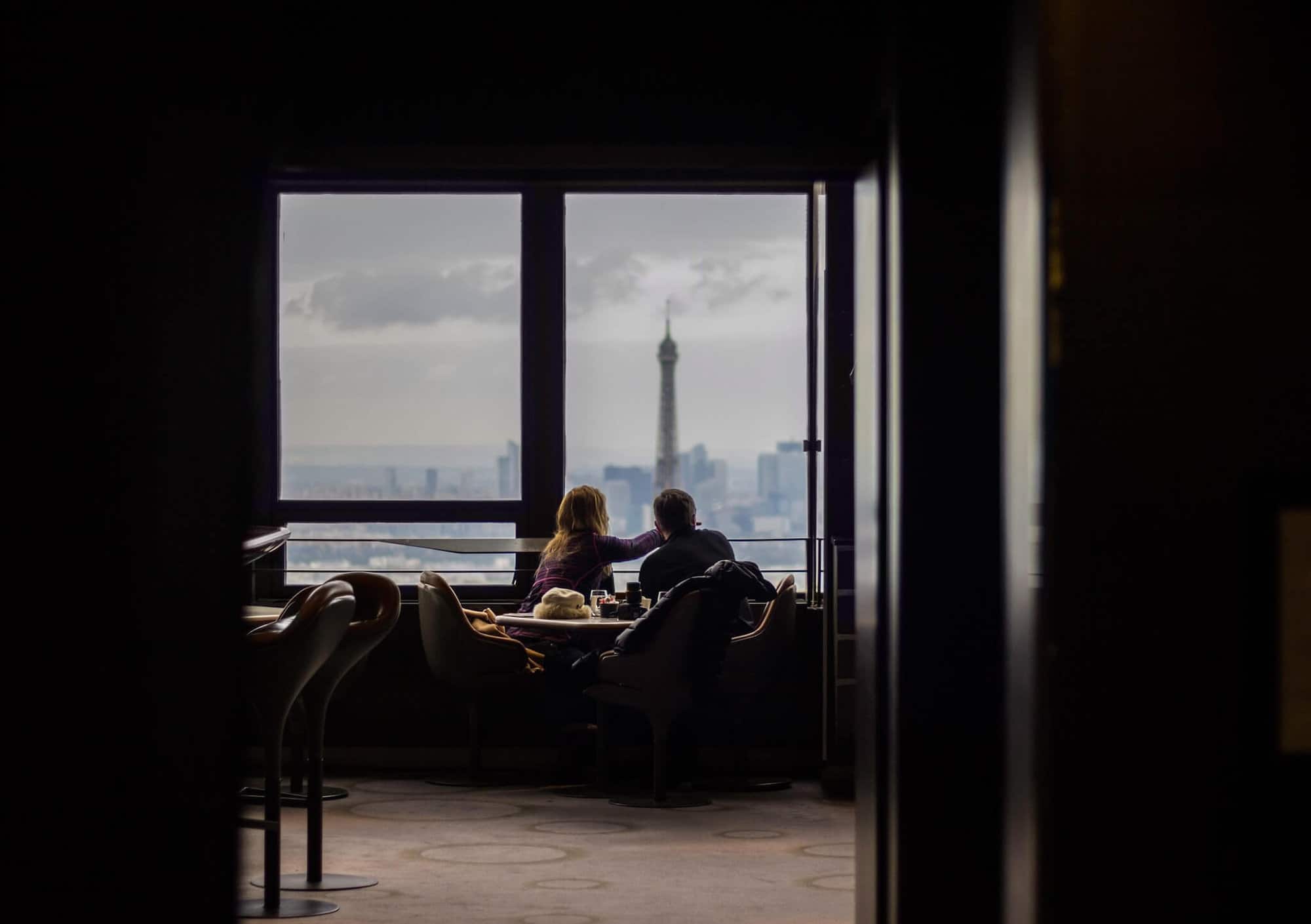 A man and a woman in a bar or restaurant look out a window where they can see Paris and the Eiffel Tower. The woman is on the left and she is pointing at the Eiffel Tower. The man is to her right.
