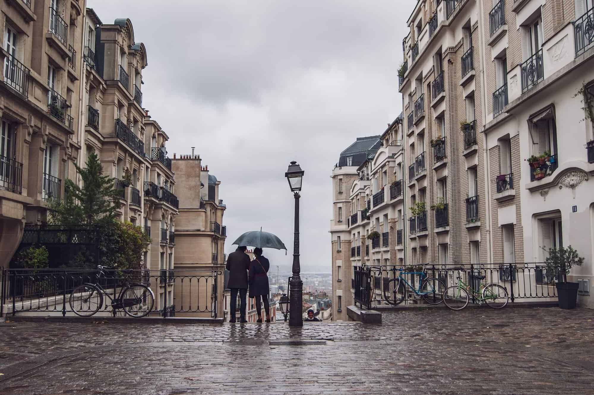A man and a woman stand under an umbrella at the top of a flight of stairs as they overlook Paris before them. There is an old fashioned street lamp to the right of the couple and there are Parisian buildings on either side of them.