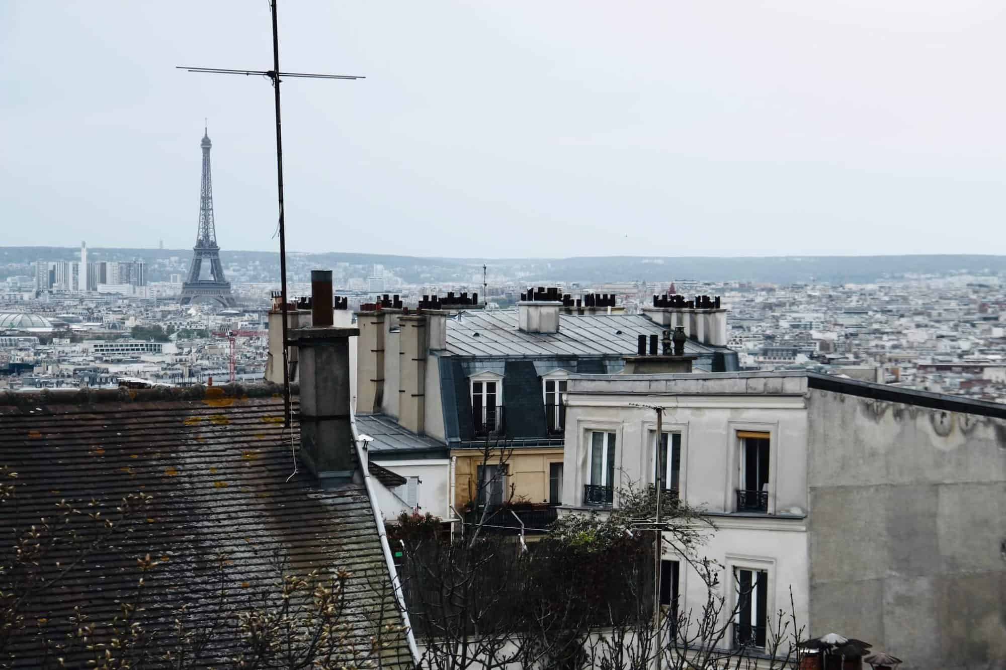 A Parisian skyline. There are several Parisian rooftops visible as well as the Eiffel Tower. The sky is gray as well as the buildings and rooftops.