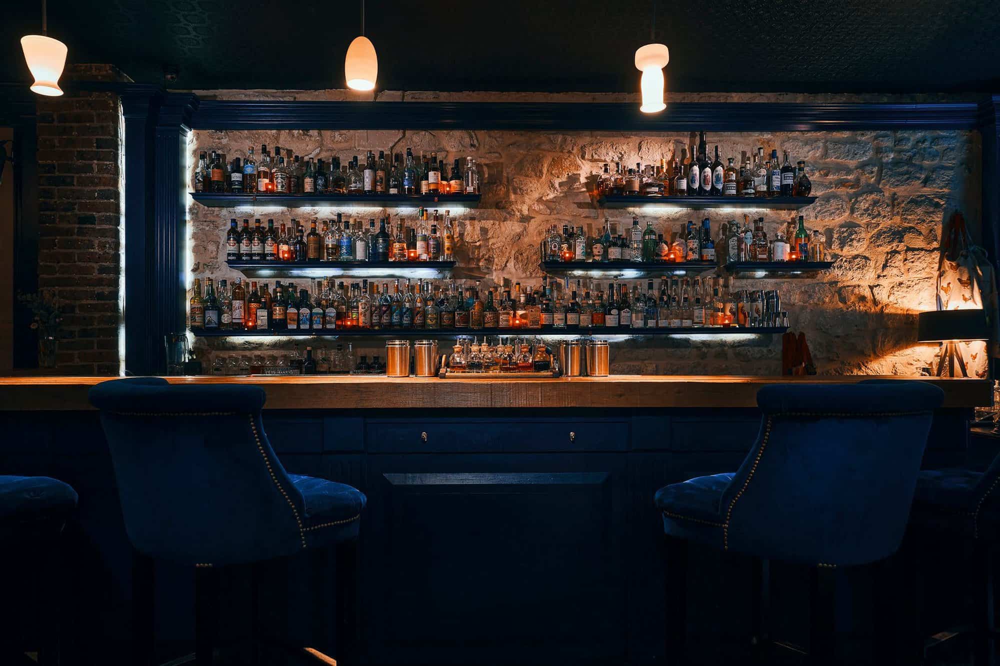 The interior of a bar in Paris called "Little Red Door." There are two high chairs covered in blue fabric at the bar and there are several bottles on several shelfs against a wall made of stone behind the bar.