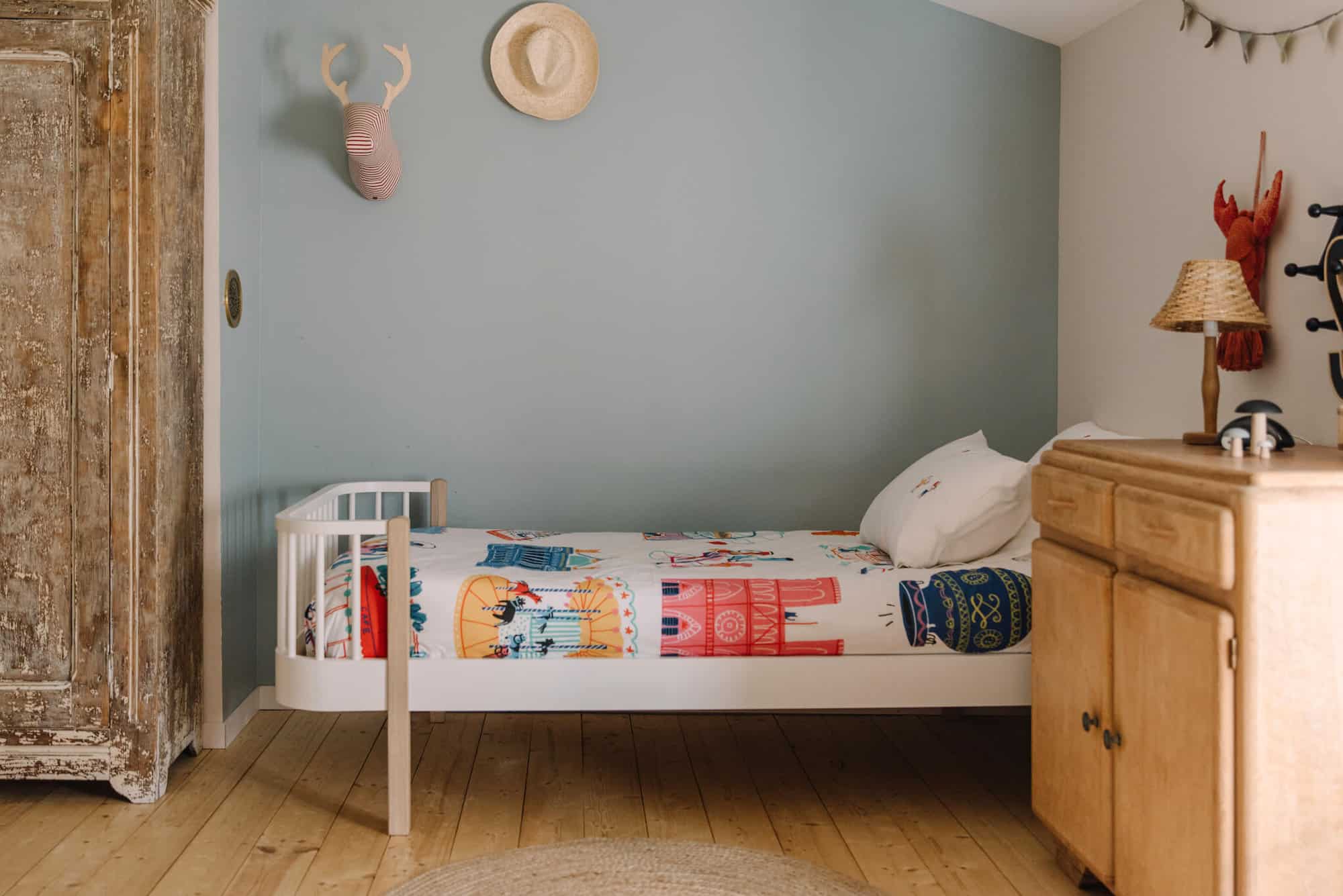 A children's bedroom is pictured, with one light blue wall and one white. The bedspread features large colorful images including a carousel and a hot air balloon.  