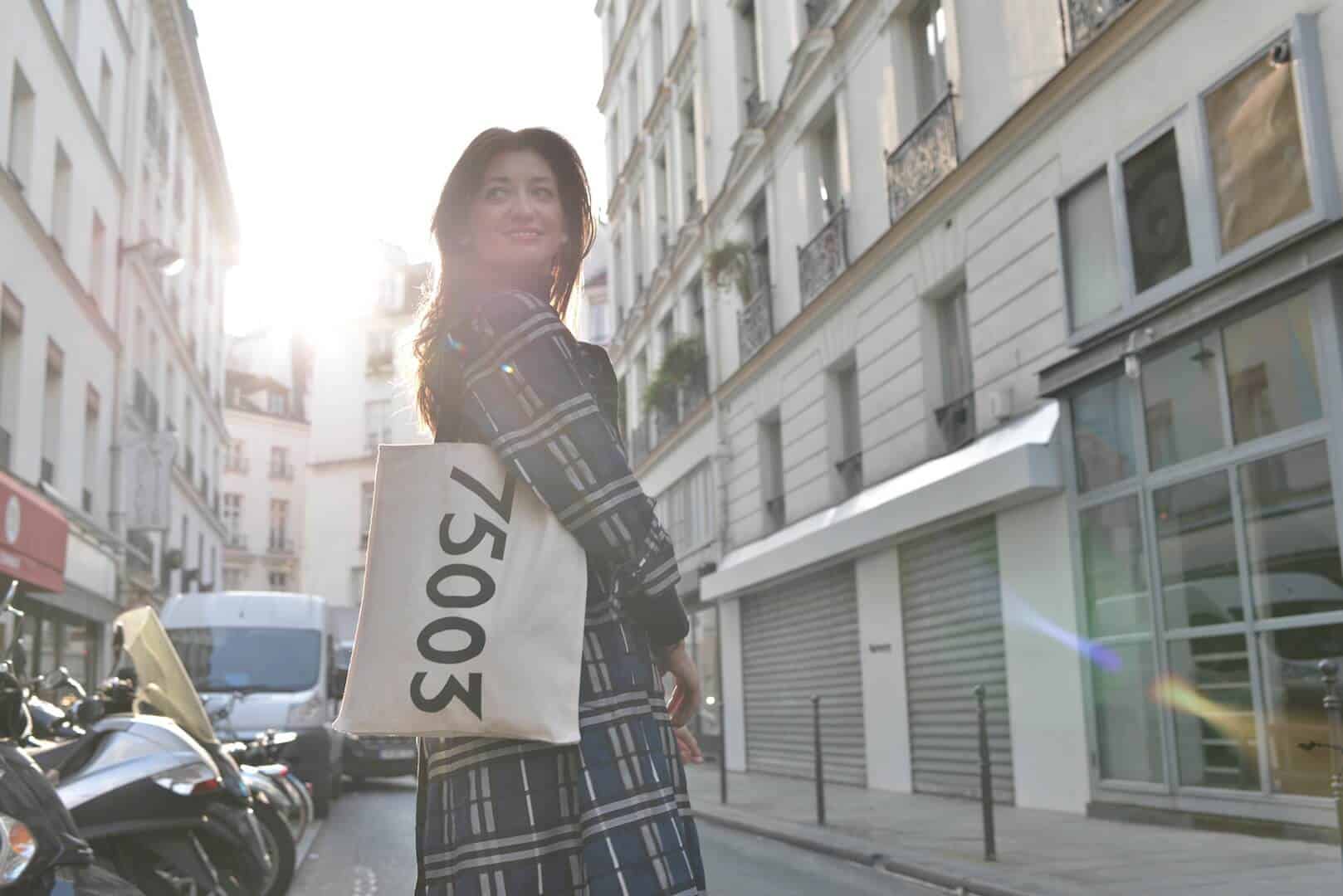A woman stands in the middle of a Parisian street wearing a striped patterned trench coat and holding a  white canvas tote bag that reads "75003" in black. 