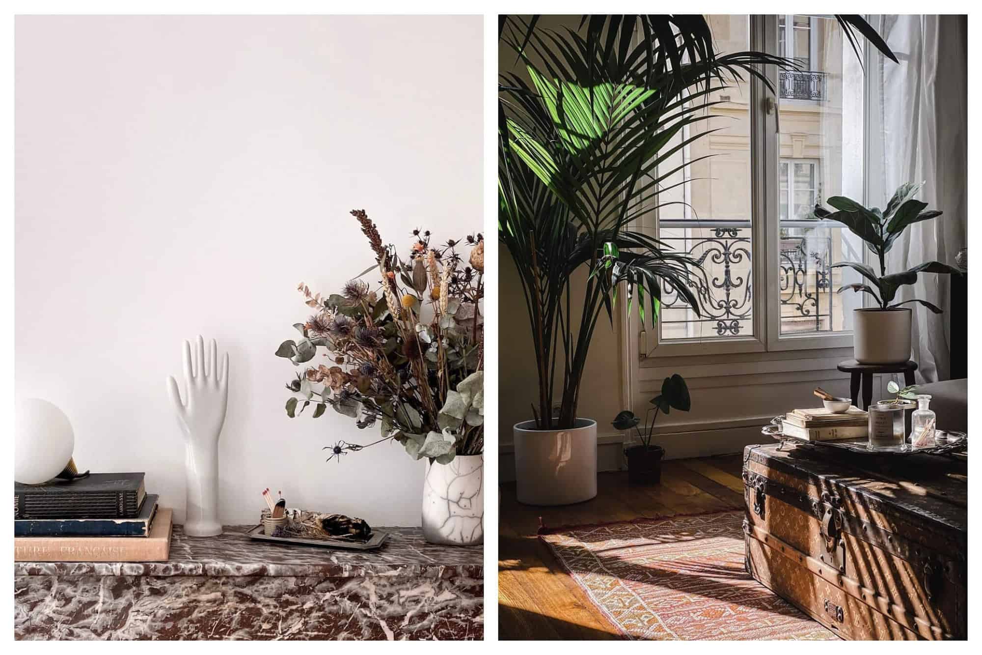 Left: A brown marble counter top is pictured with a variety of accessories on top including books, a ceramic hand and wrist sculpture, matches and a vase with dried flowers. Right: A sun-filled room within apartment is pictured with two large potted plants. There is a Louis Vuitton trunk used as a coffee table and a large window with a Haussmanian balcony and a view of the exterior of a Parisian apartment.
