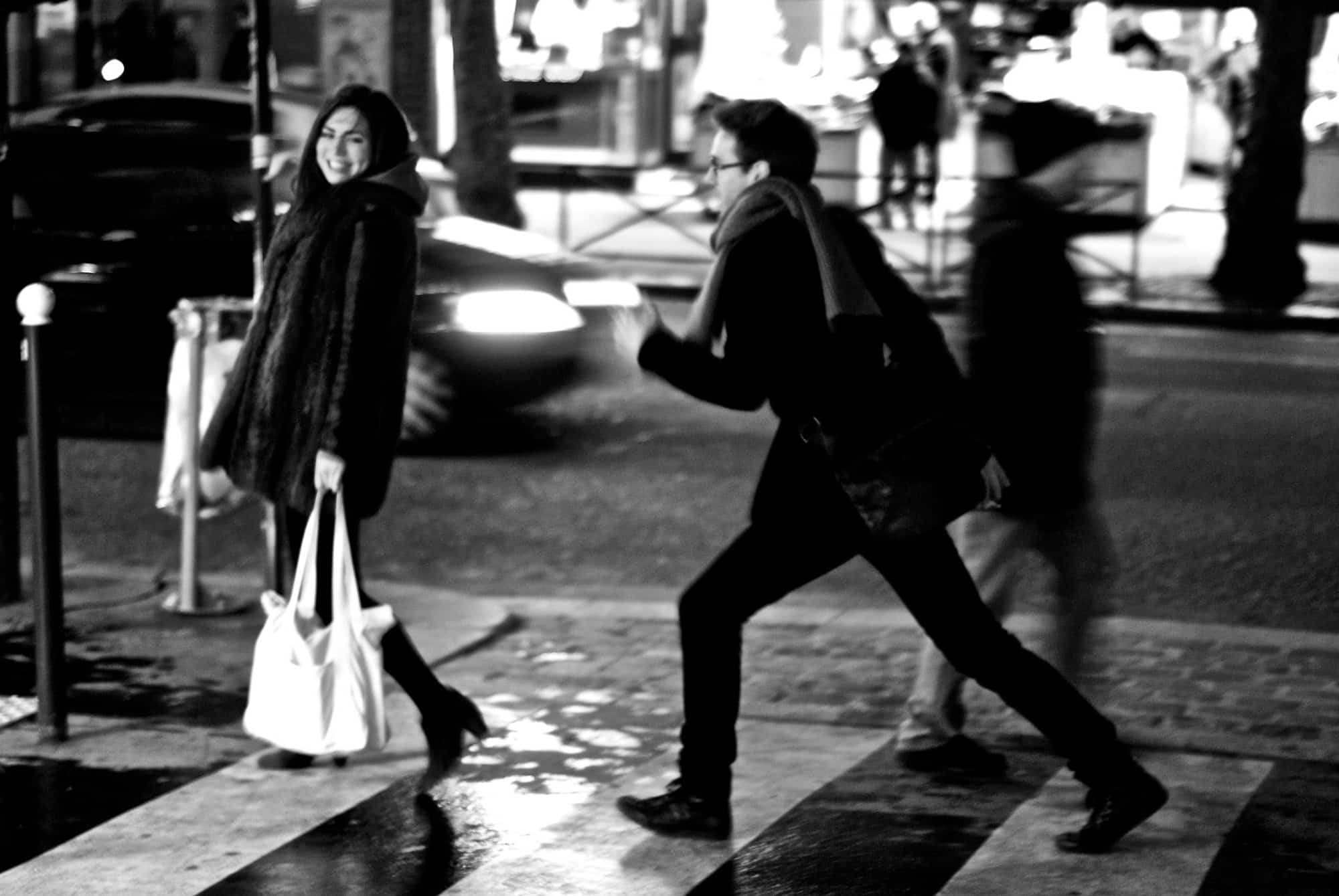 A man and a woman are crossing the street with the woman smiling playfully and the man appearing to run after her.
