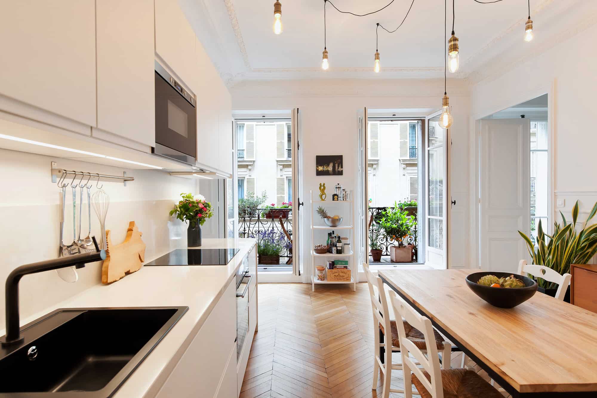A picture of a spacious Parisian apartment. In this photo taken from an American-styled open kitchen, you will see 2 tall french windows opening to the balcony full of plants. The kitchen is sophisticatedly decorated in white and tan. 