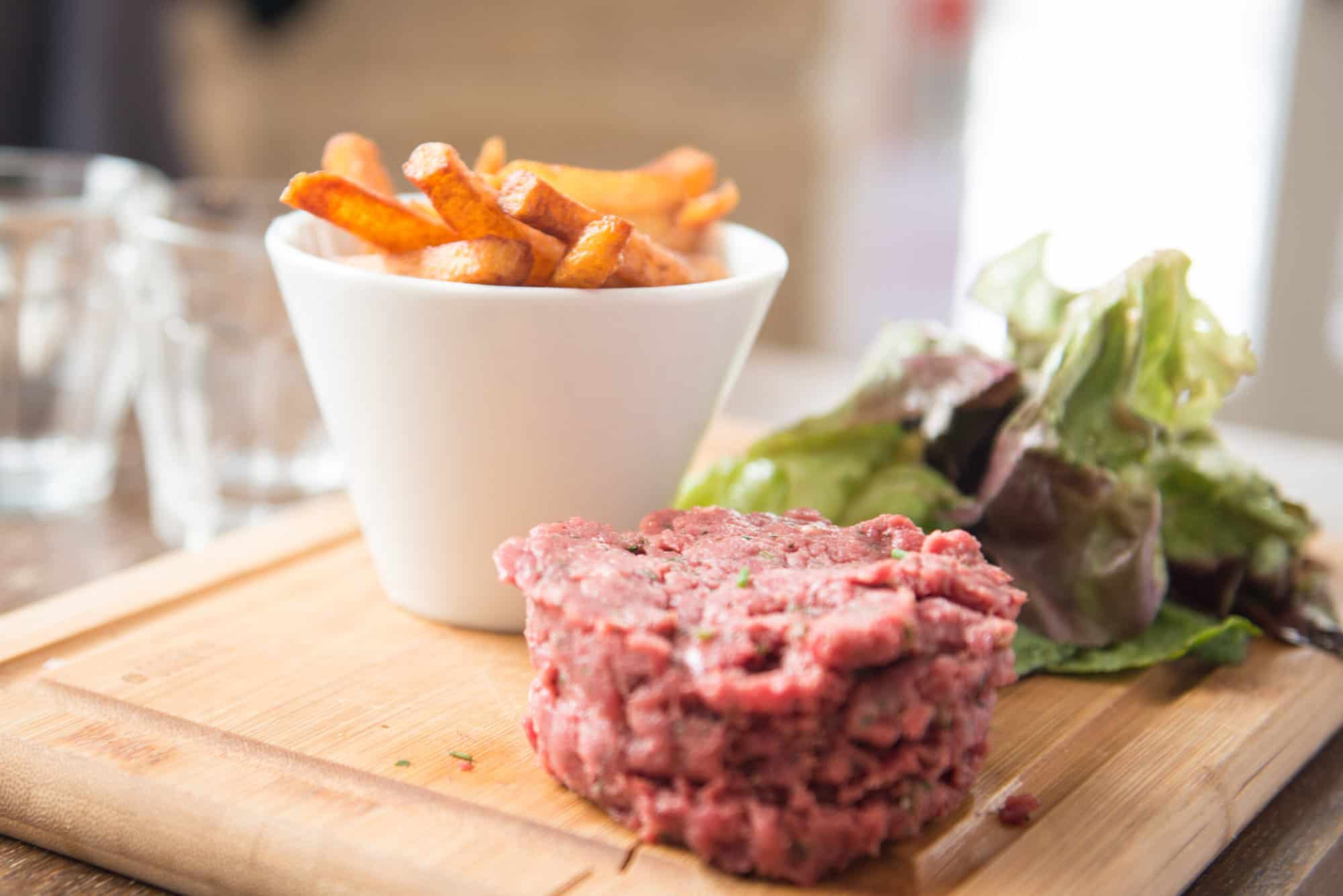 A close up of one of their menu, the Steak Tartare, served in a wooden board with salad and fries on the side. The fries is served in a white small cylindrical bowl.
