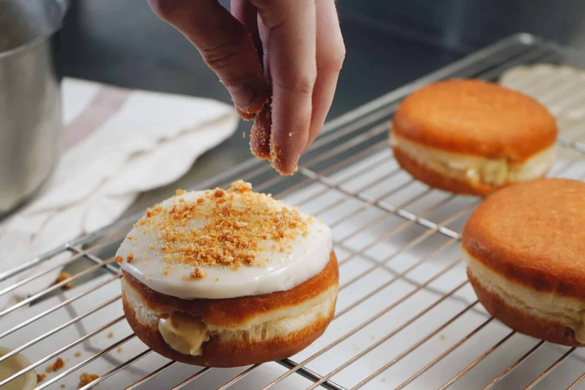 An iced, filled donut on a cooling rack while a person sprinkles crunchy speculoos over the top along with two un-iced donuts cooling on the rack as well.