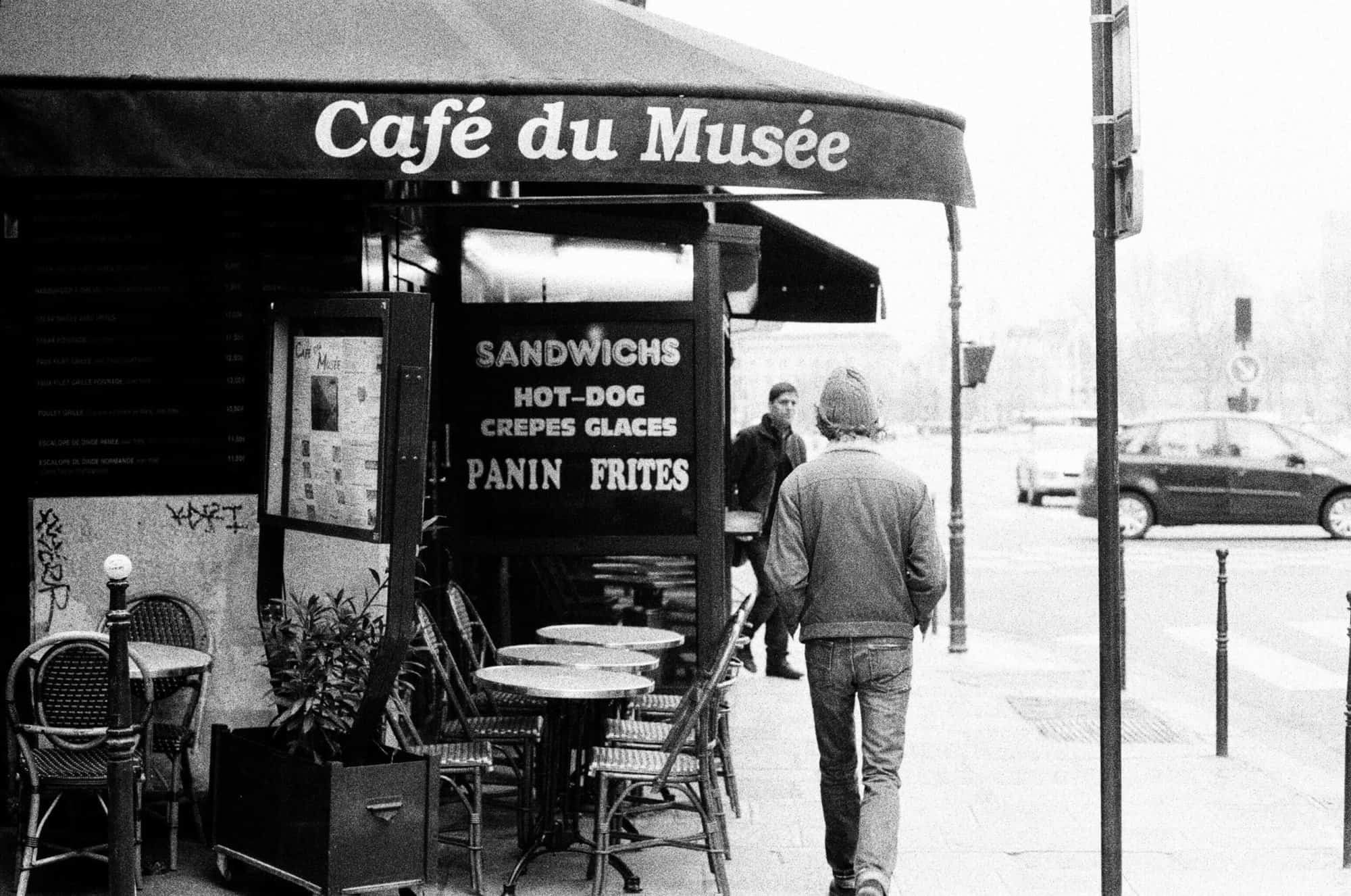A Parisian café and people walking in front of it. The sign on the café reads Café du Musée. 