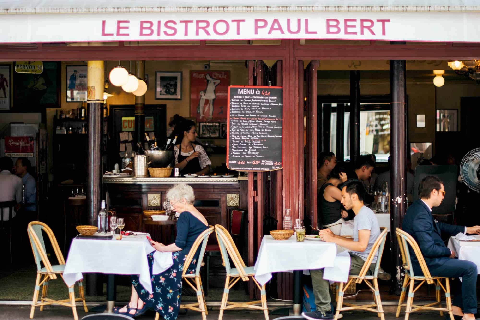 The front of le bistrot paul bert with cane chairs and tables with white table cloth. There are also people dining. 