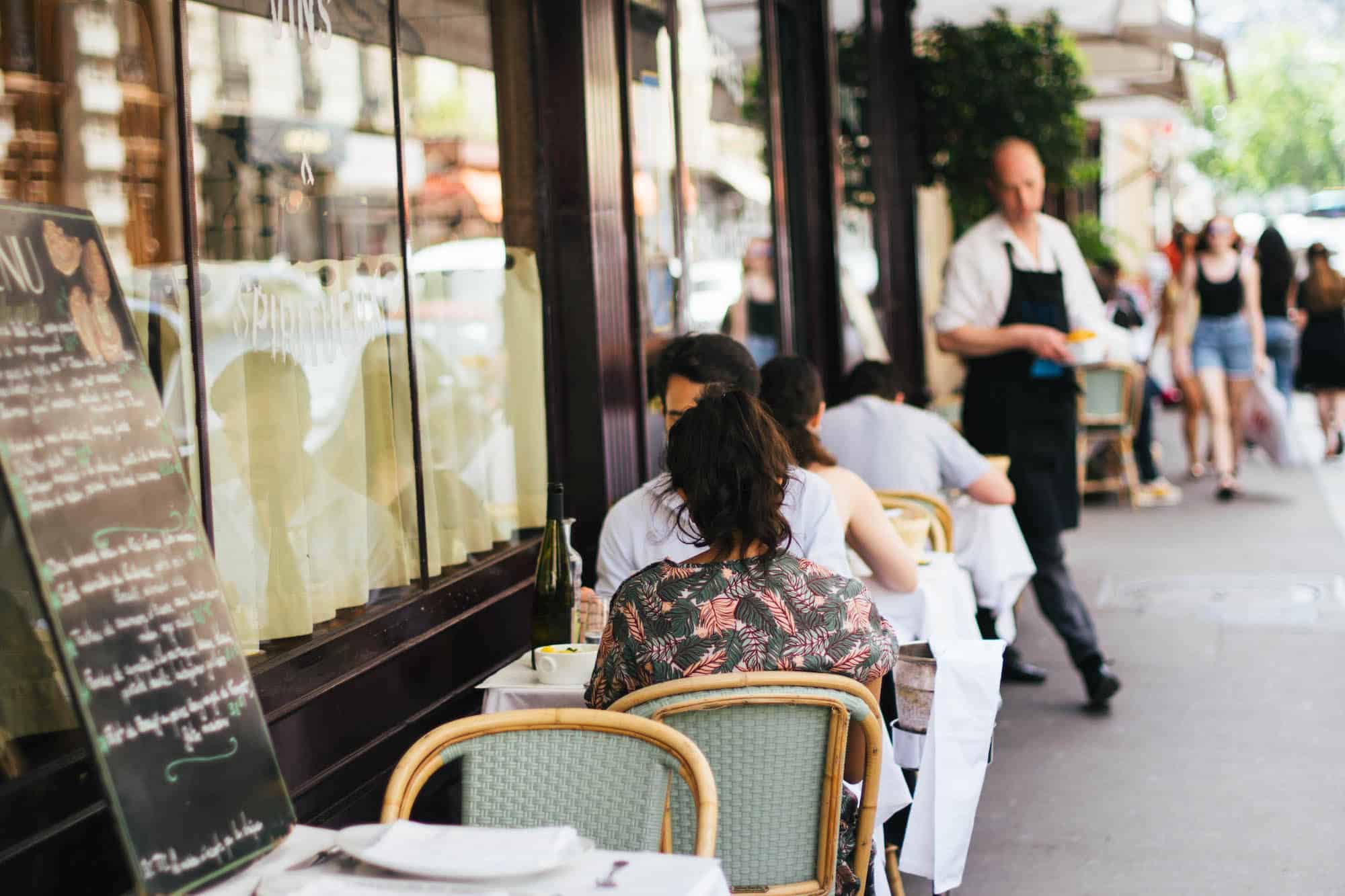 A couple sitting on the terrace of the Paris restaurant Bistrot Paul Bert. There is a chalk board menu and a waiter in a black apron coming out to serve them, with people walking down the street in the background.