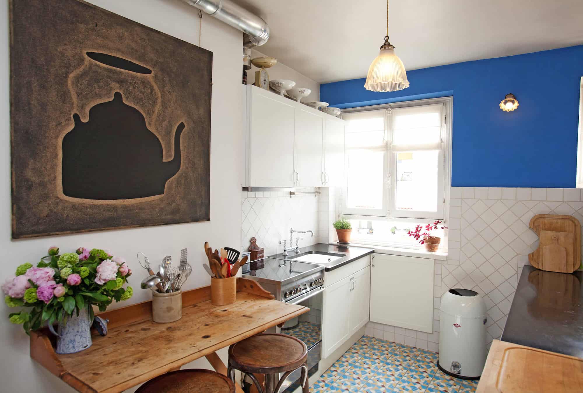The kitchen of a Parisian apartment with patterned tiles, a blue feature wall, a wood breakfast bench with flowers, and a large artwork on the wall. 