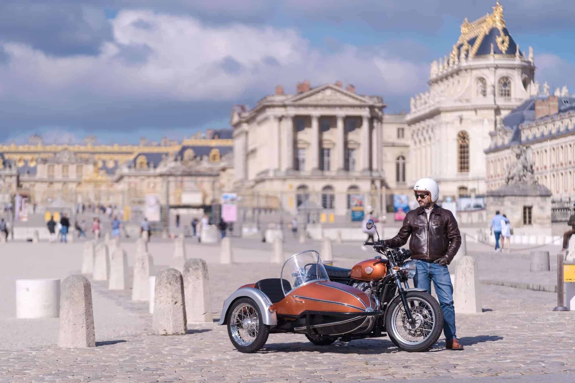 Simon Burke standing next to his vintage motorcyle in front of Château de Versailles.