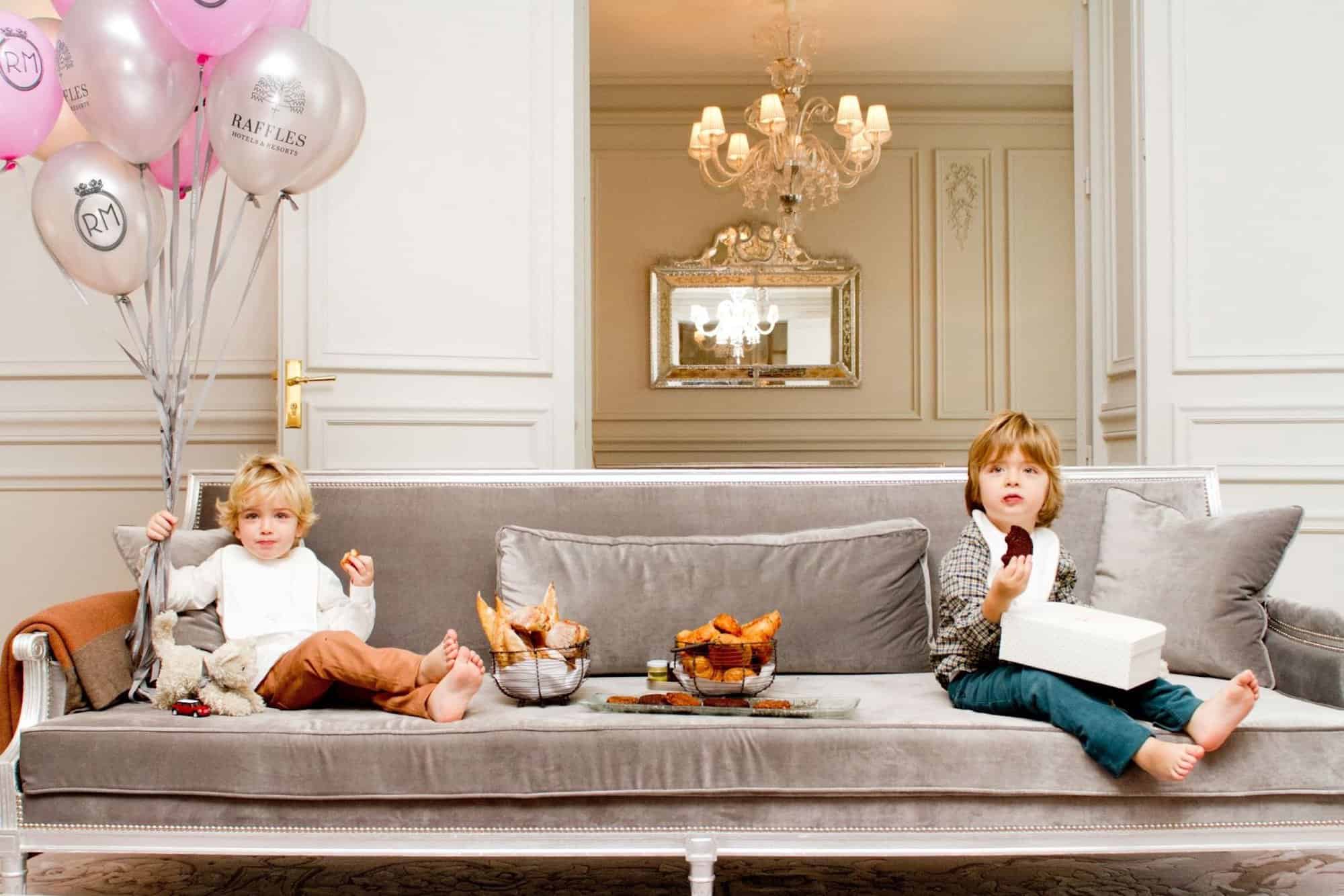2 young boys sit on a sofa in a hotel room, with balloons. they are enjoying cookies and croissants. 
