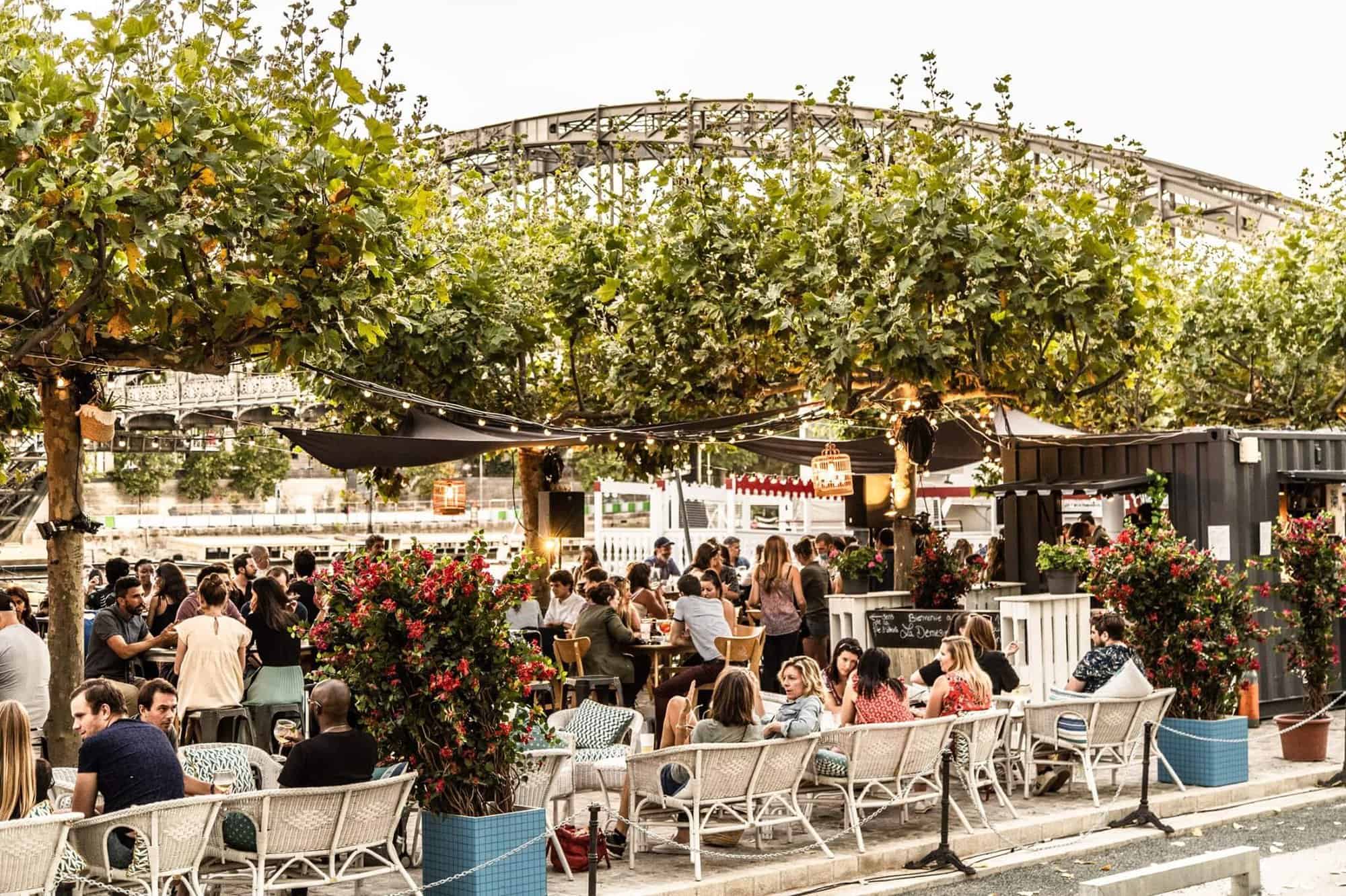 A bustling terrace next to the Seine, shaded by trees and surrounded by plant pots.