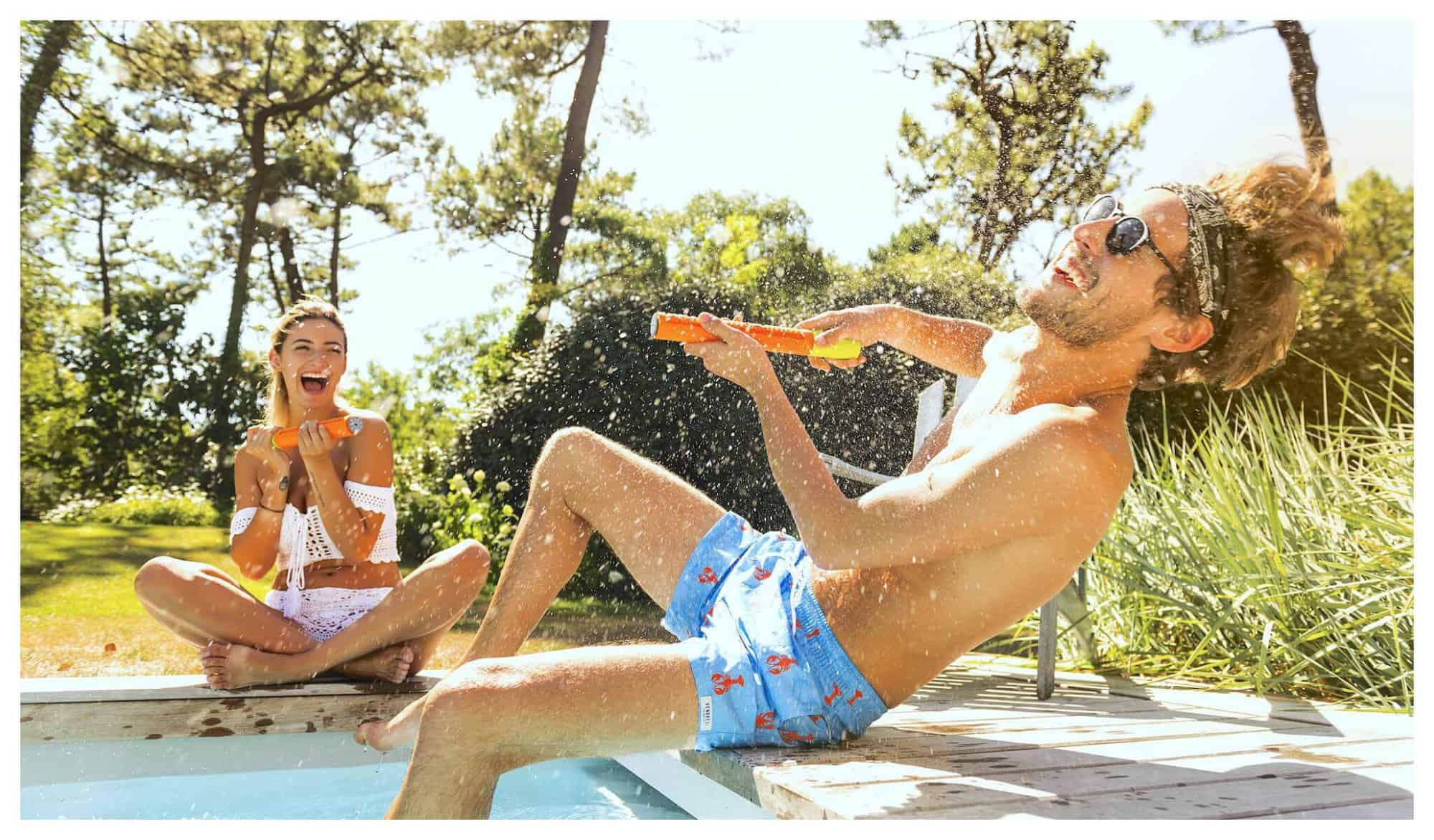 A man and a woman by a pool are having fun splashing water to each other with orange water guns.
