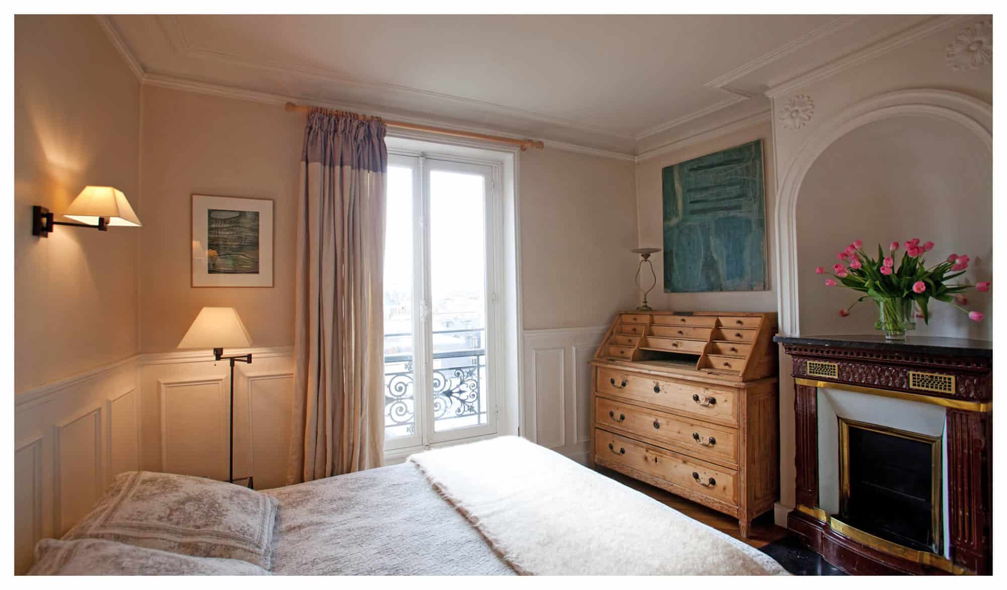 The bedroom in Erica Berman's apartment with white and brown sheets, a wooden commode, a bouquet of pink tulips, and 2 white wall lamps.