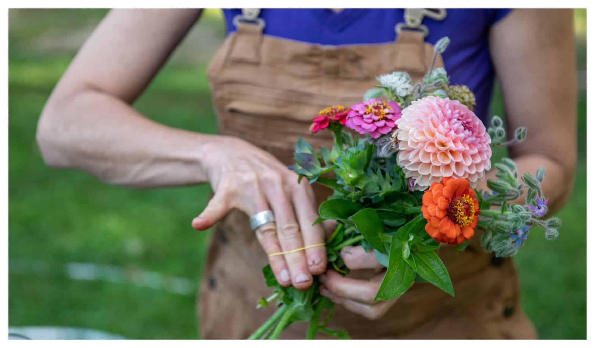 A woman is holding a bouquet of orange, pink, and red flowers from the Veggies to Table farm.