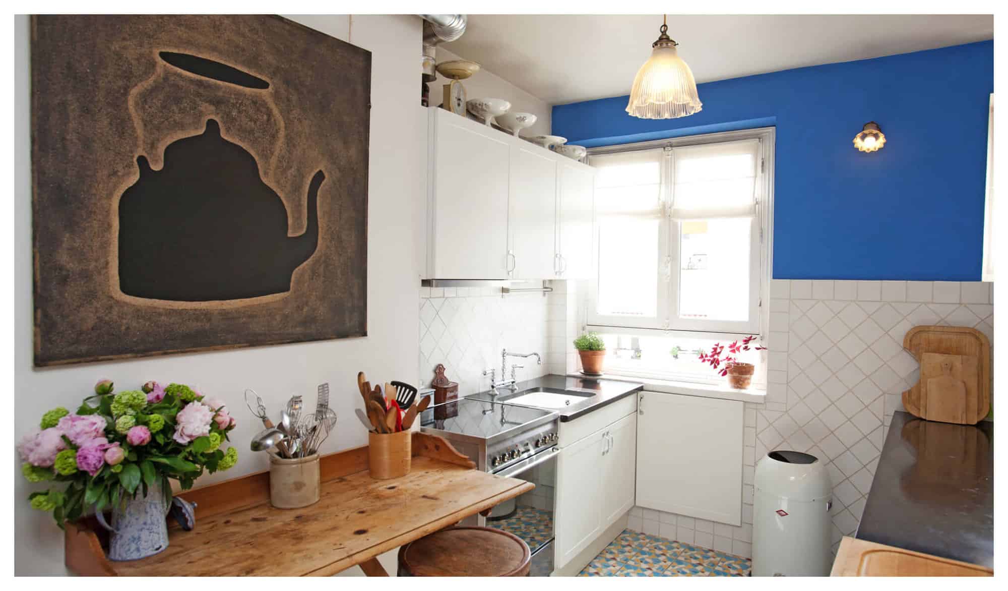 The kitchen of Erica Berman's Parisian apartment, with white cabinets, blue walls, wooden table, and a black kettle artwork.