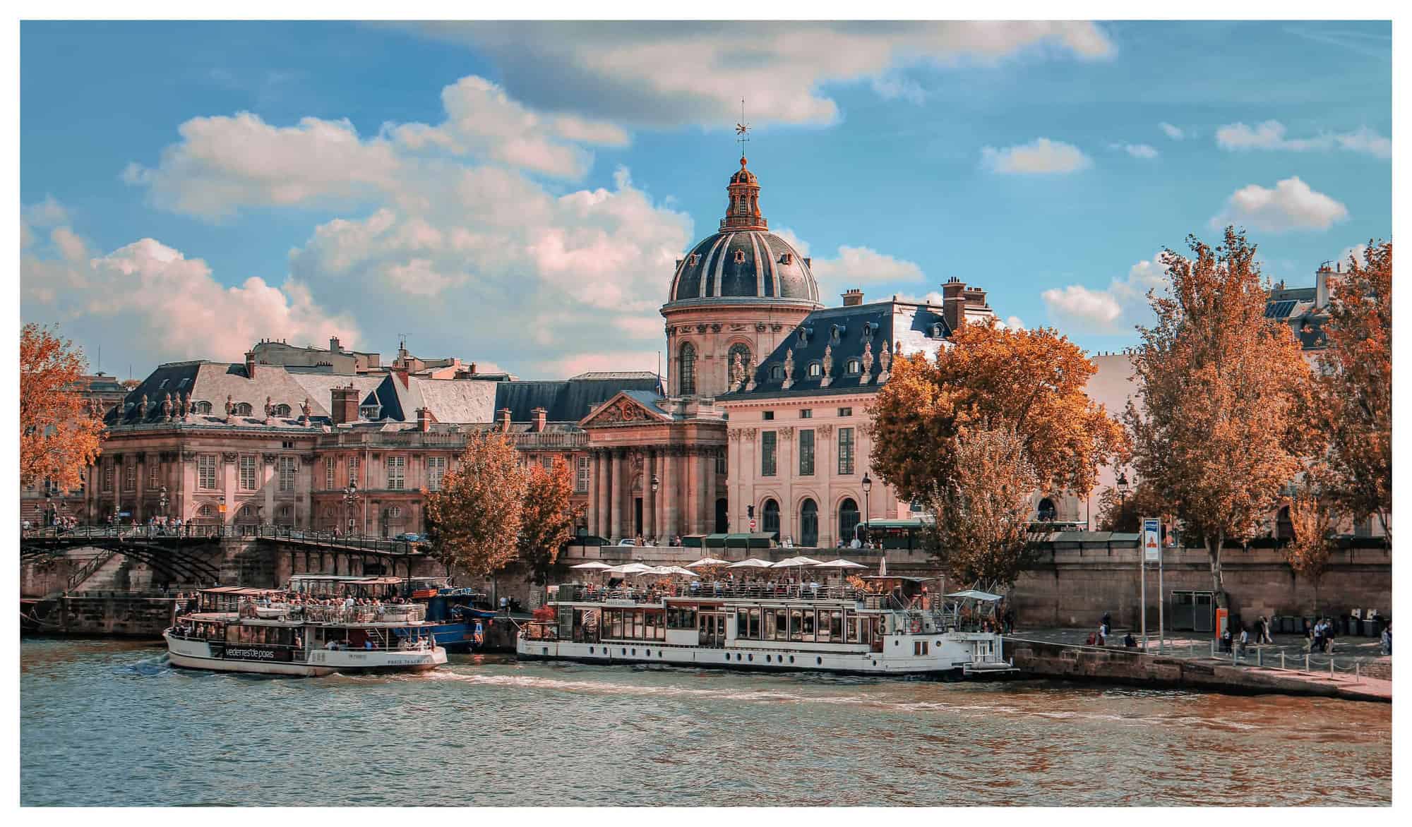 A view of the Seine river in the middle of an autumn day, with white boats, brown leaved trees, and rustic buildings.