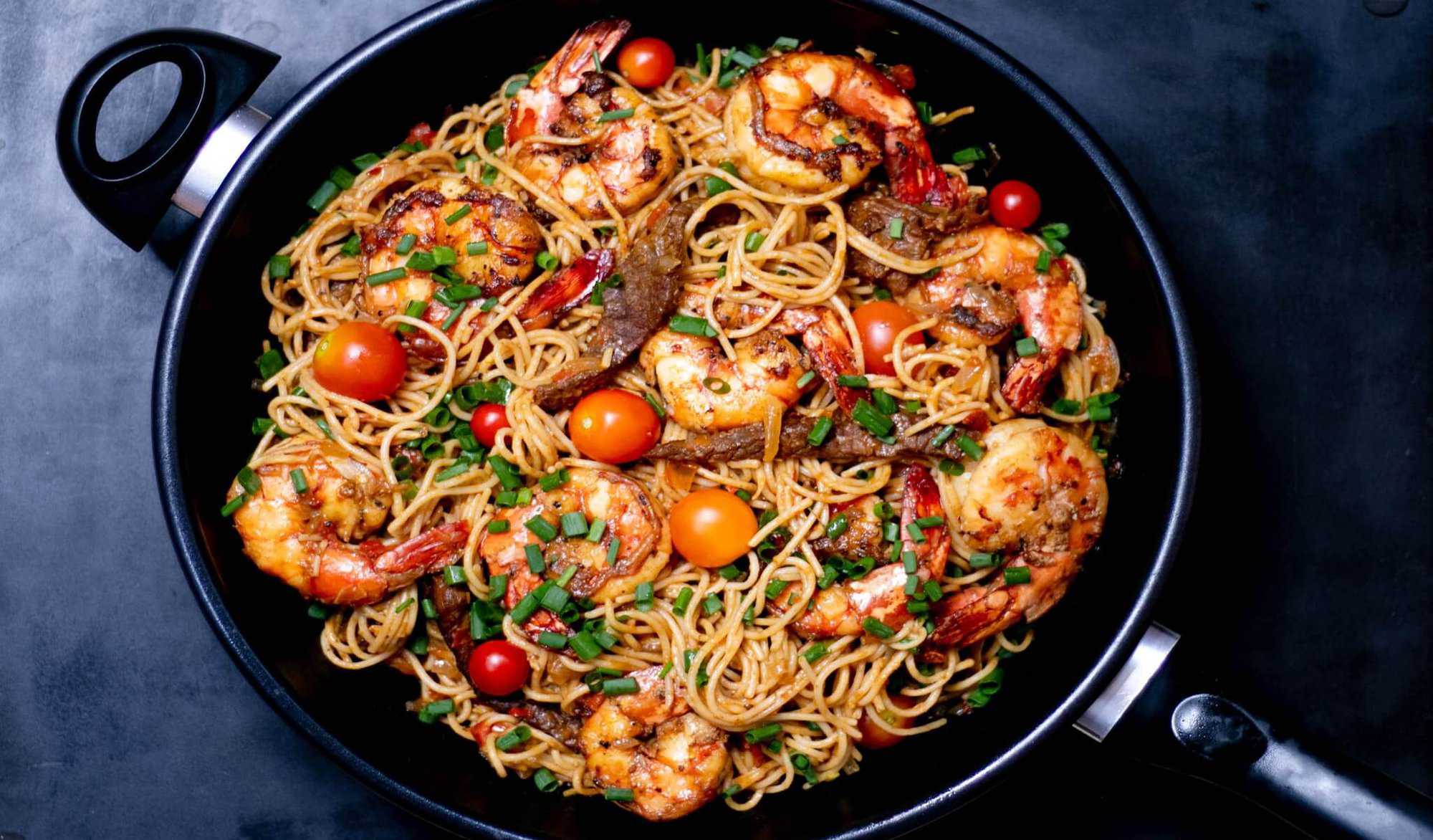 A pan full of seafood pasta with cherry tomatoes, grilled prawns, and chopped herbs.