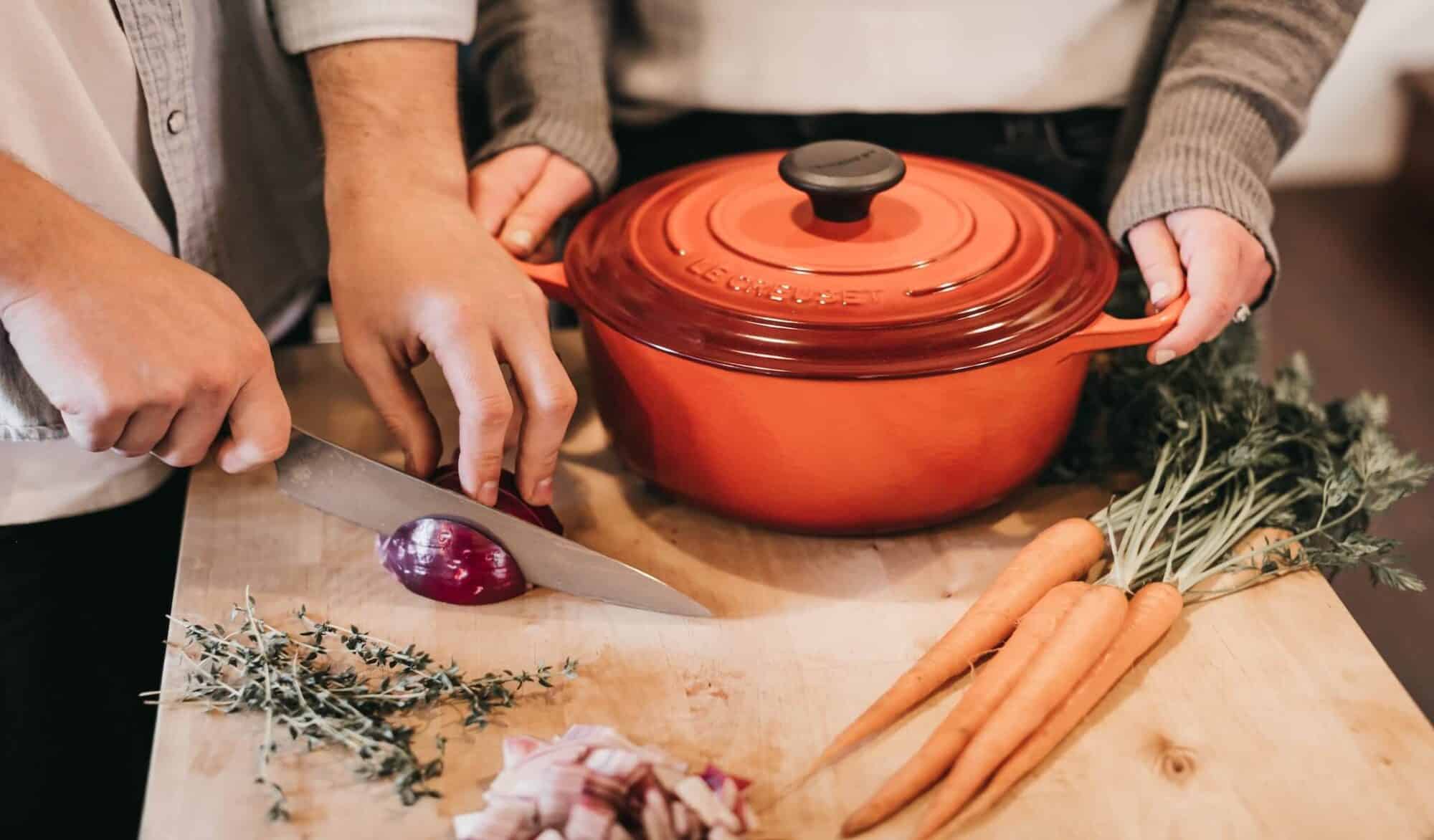 A man chopping a red onion with a long knife while a woman holds a red oval dutch casserole 