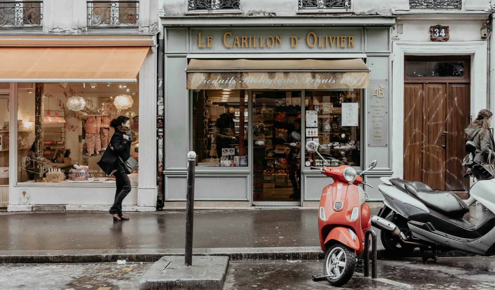 A woman in an all-black outfit walks by a Parisian pavement with stores and a parked red Vespa motorcycle
