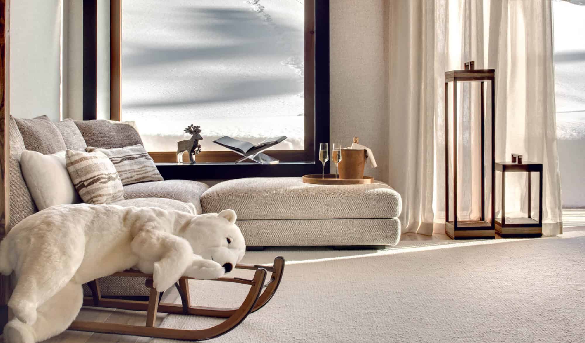 A living room at Antarès resort, with a beige couch, a large polar bear stuffed toy, and two glasses of champagne all situated next to a large window looking onto the snowy slopes.