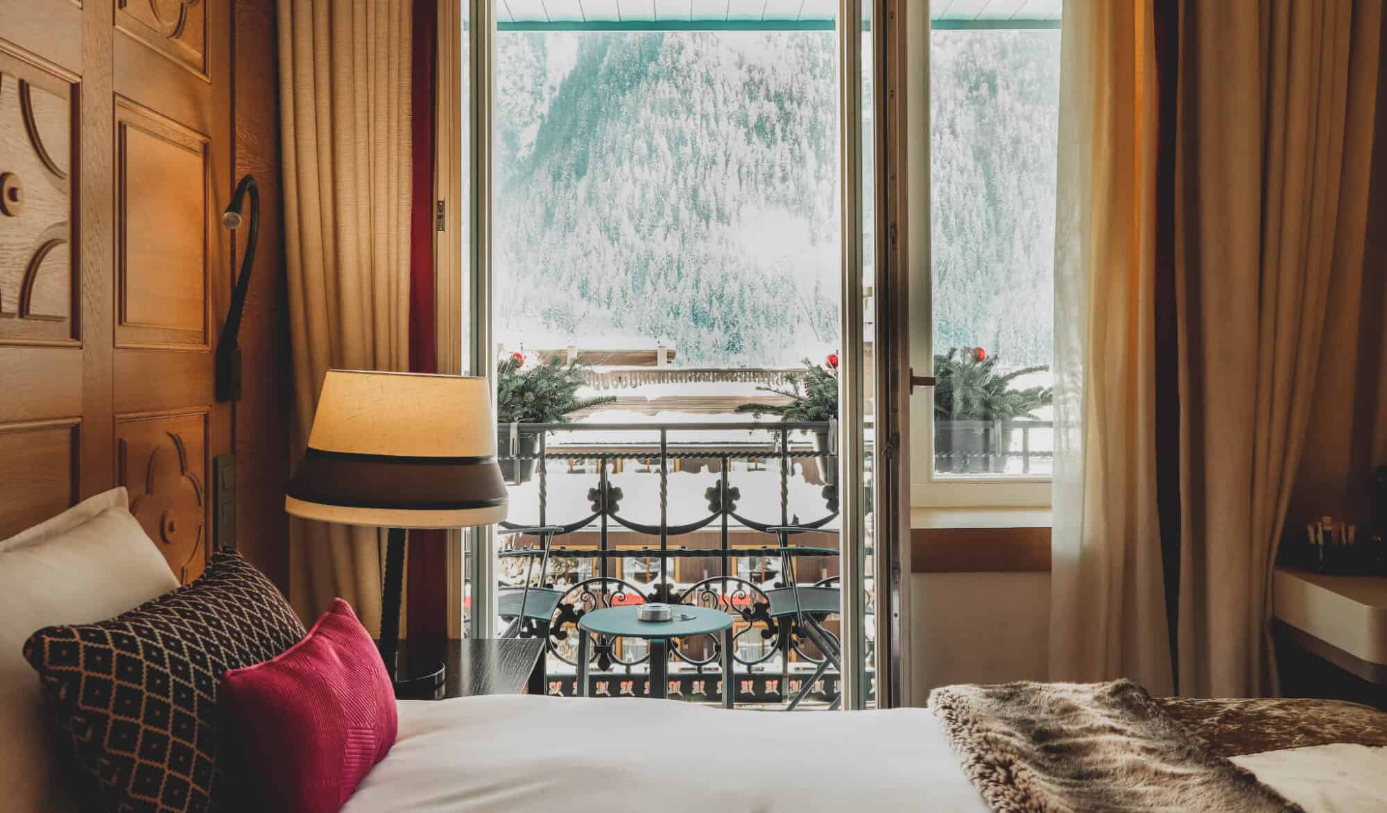 A hotel room at Hotel-Mont Blanc, Chamonix with a bed against a wood paneled wall, white bedding, beige, burgundy and red pillows, and a view of a snowy mountain slope.