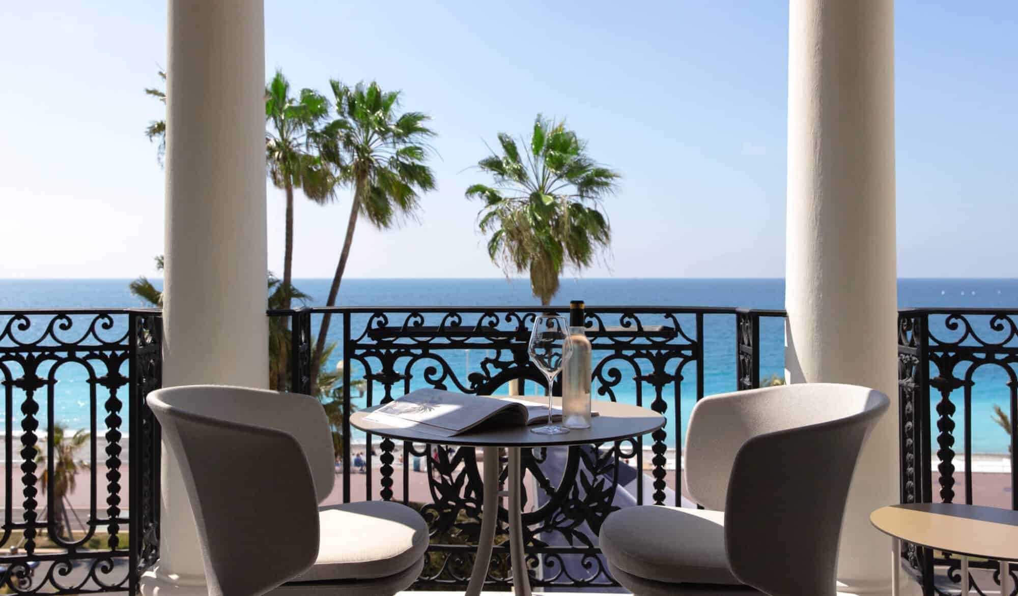 A sunny balcony of a suite from L'Hotel Negresco with a view of the beach and a few palm trees. There sits a table for two on the balcony with a black iron railing, a book and a bottle of wine waiting for an occupant.