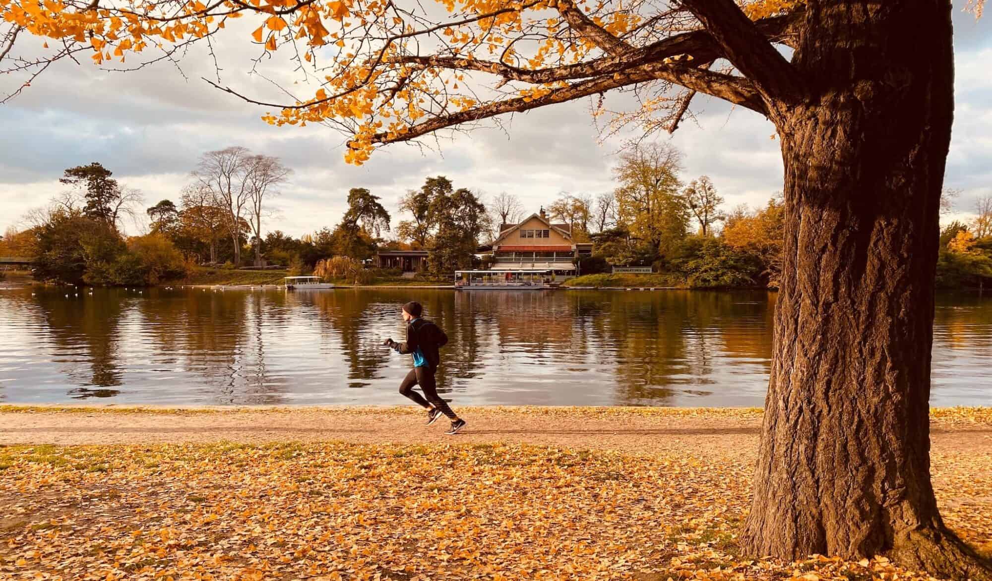 A jogger dressed from head to toe in black running gear jogs along a pond in the Bois de Boulogne in Paris during Autumn.