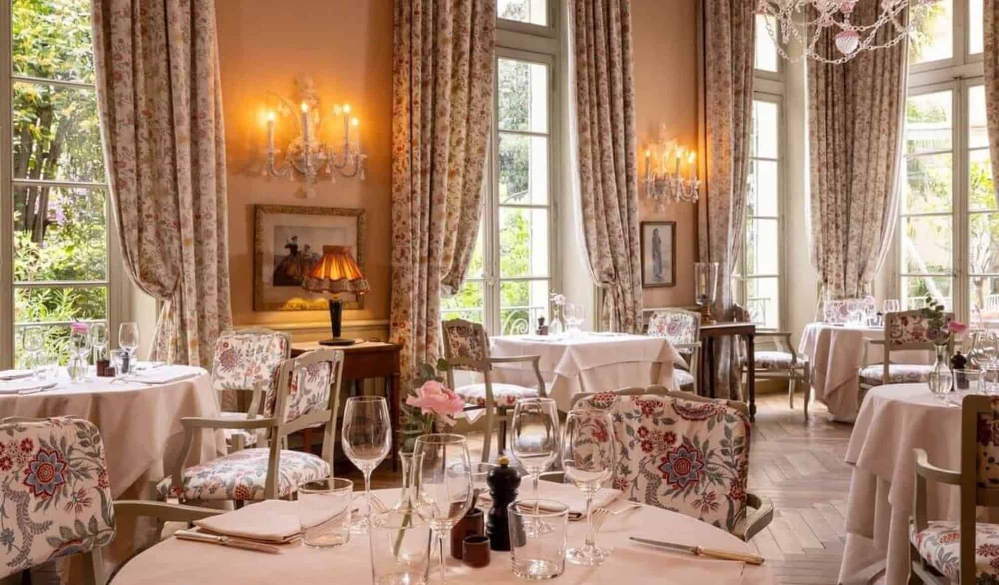 The breakfast room in La Mirande Avignon decorated in dusty rose with floral patterned curtains and upholstery, large windows and parquet flooring.