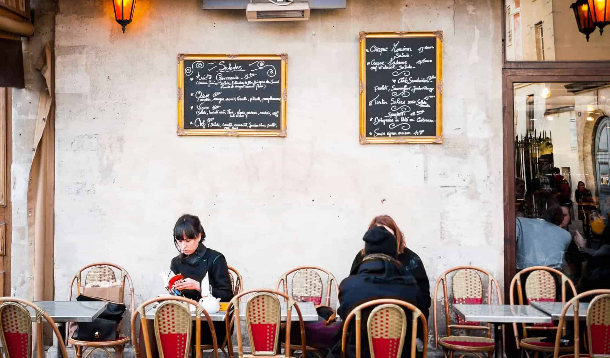 A terrace of a Parisian restaurant with 3 people sitting in red and brown rattan chairs, with menus on slate boards posted on the stone wall.