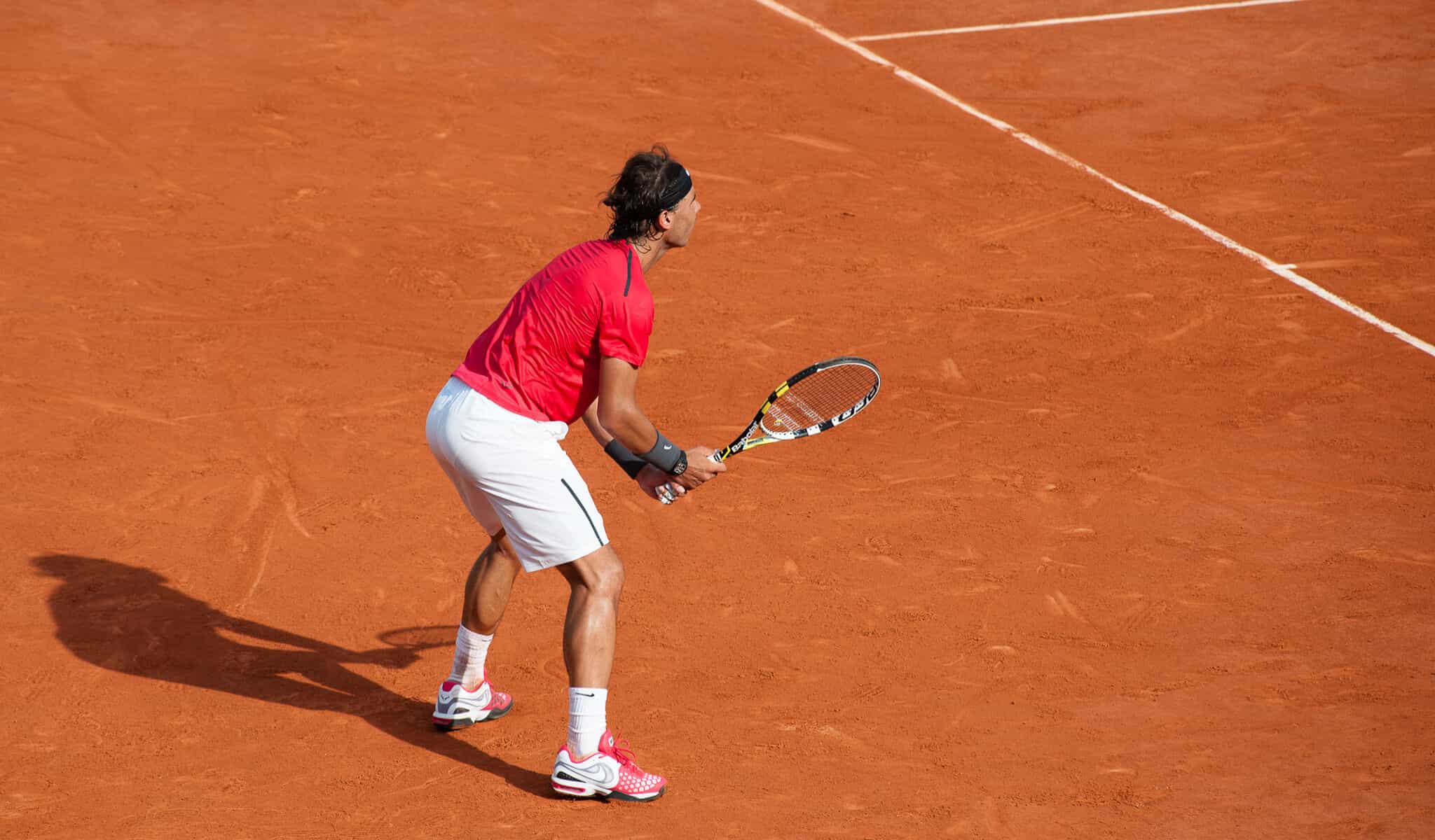 Rafal Nadal plays at the French open in red shirt and white shorts, on the iconic orange clay courts.
