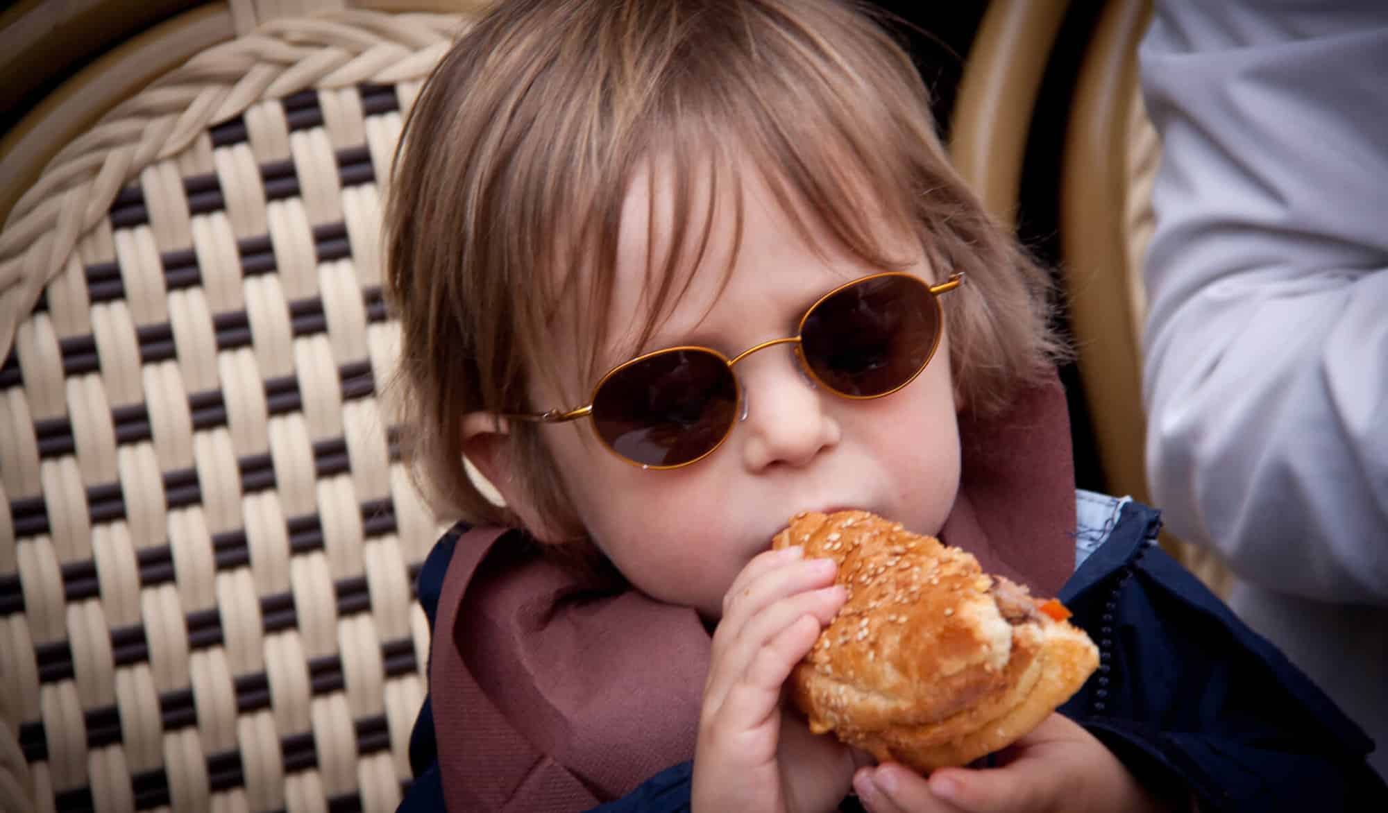 A French kid with golden brown hair and sunglasses takes a bite from a burger he holds with two hands.