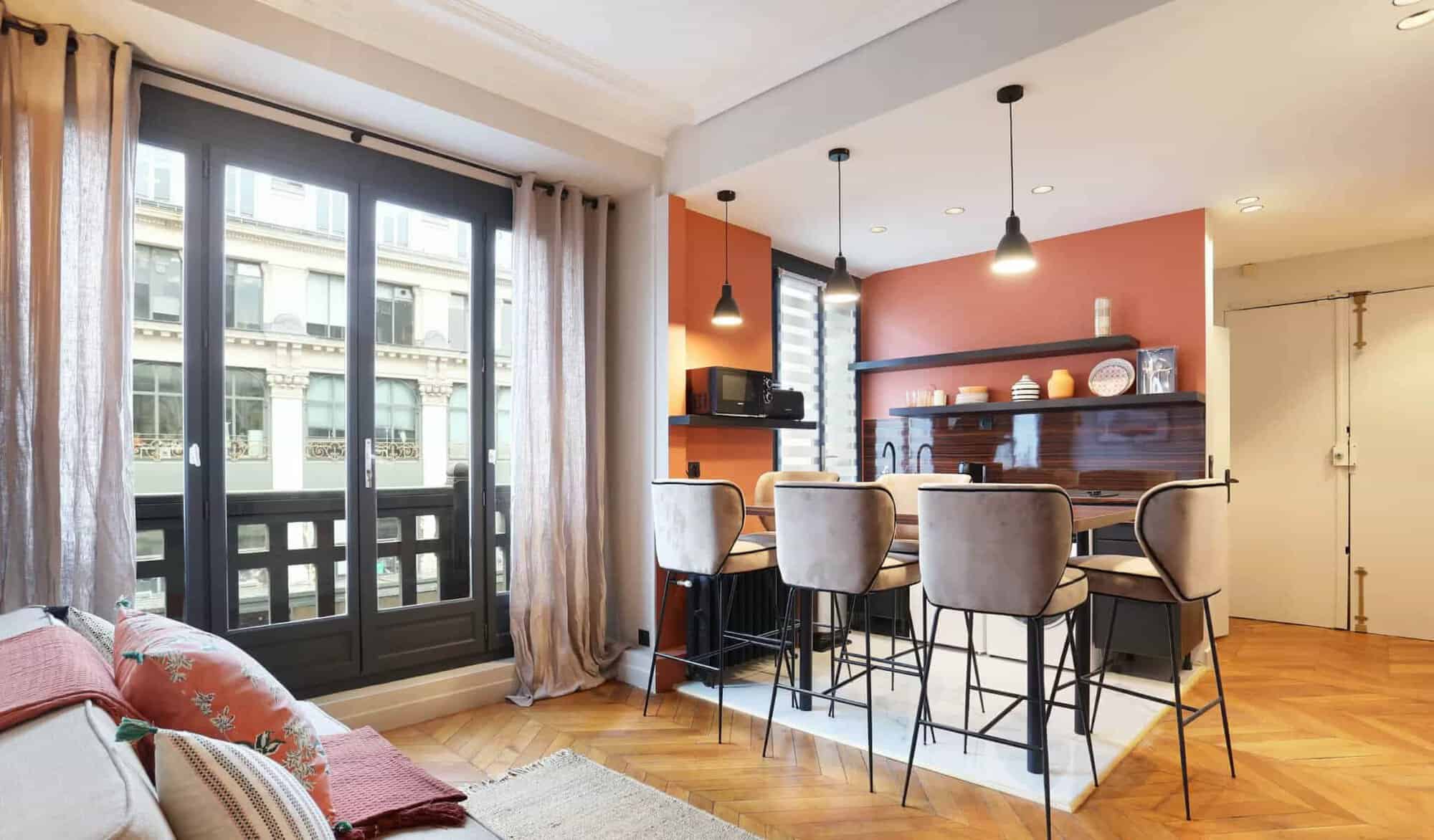 A modern living room and kitchen with orange walls and decorated with black and brown lights, gray bar stools, and beige curtains.