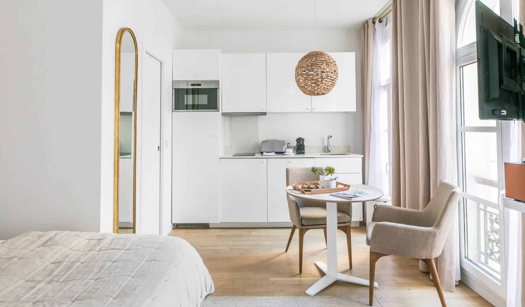 A studio with white walls and kitchen cabinets, a bed with beige linens, beige curtains, a white round dining table and 2 beige chairs.