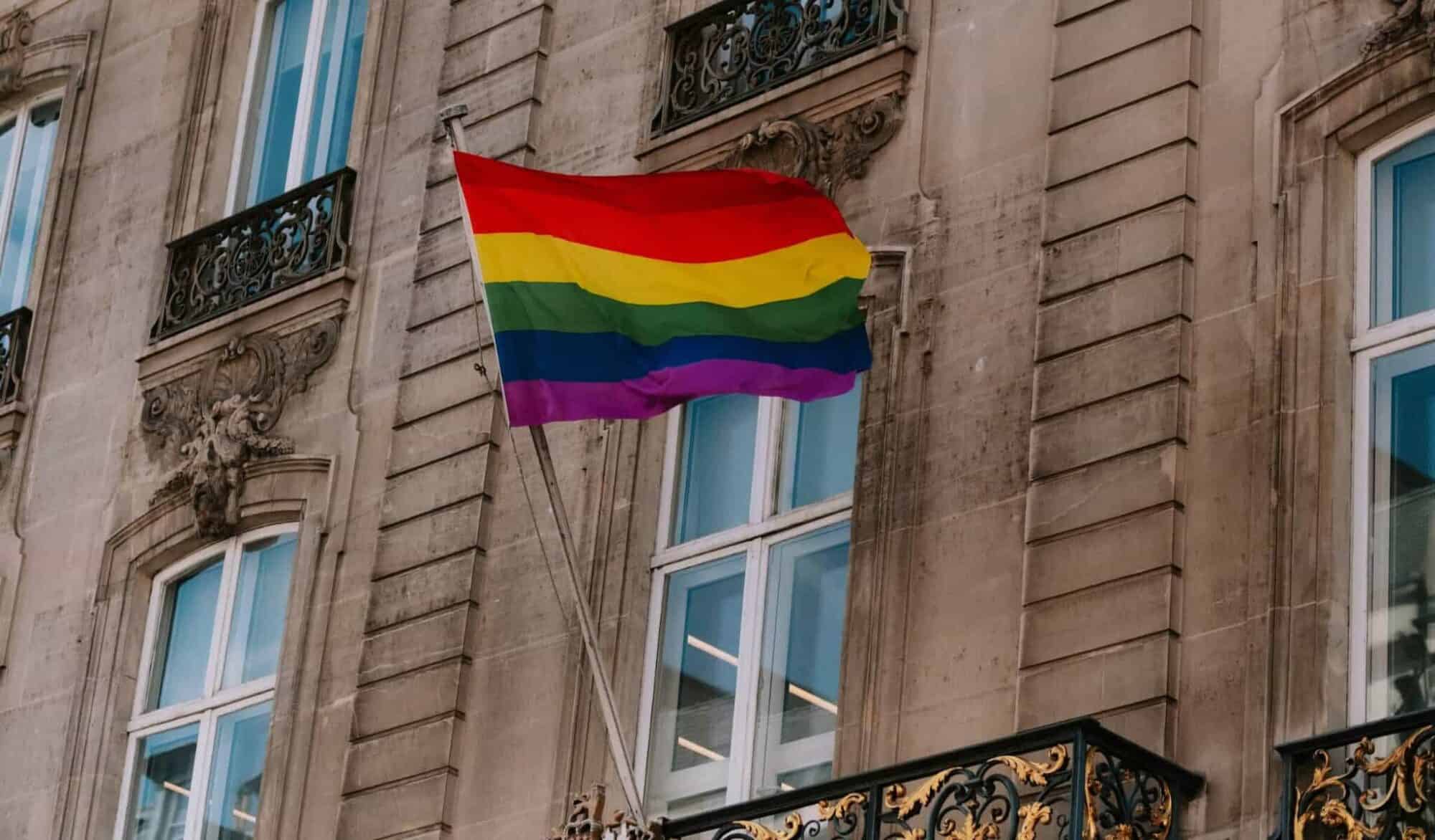 The LGBTQ+ rainbow flag waves in front of a Parisian window.
