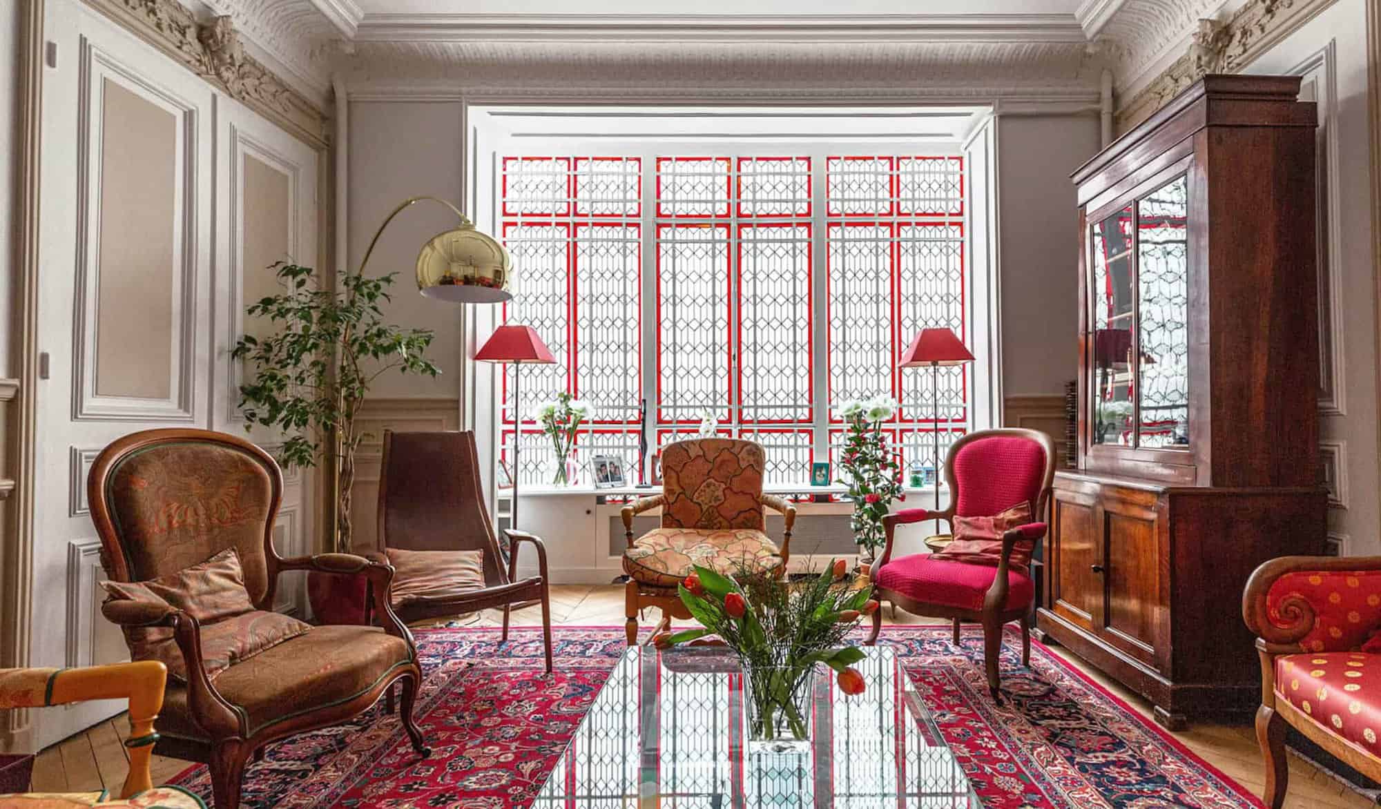 A Parisian living room with red windows, red Persian carpet, wooden chairs with red cushions, and a tall wooden cabinet.