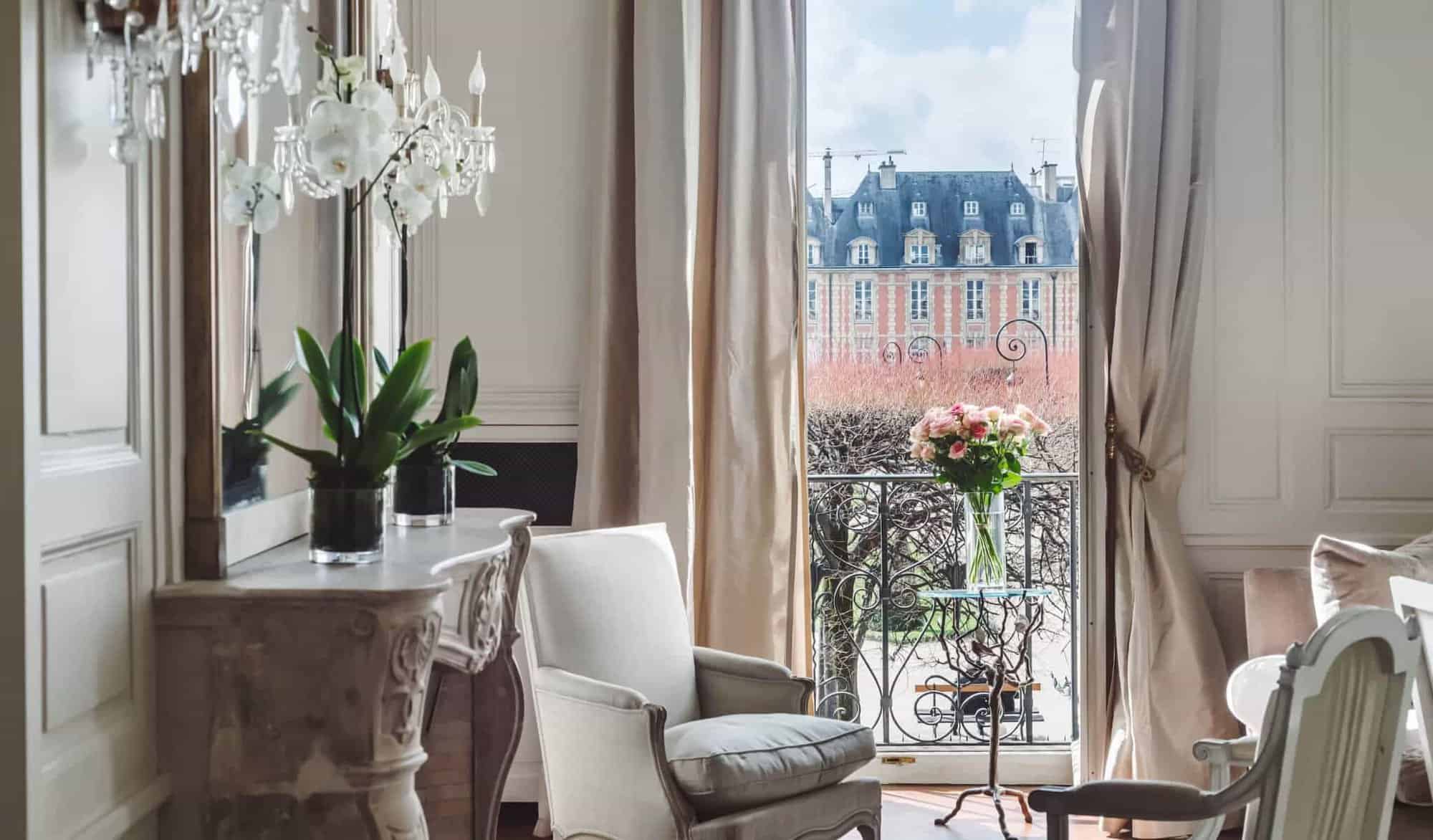 A Parisian living room with a window and balcony overlooking the Place des Vosges iconic orange buildings.