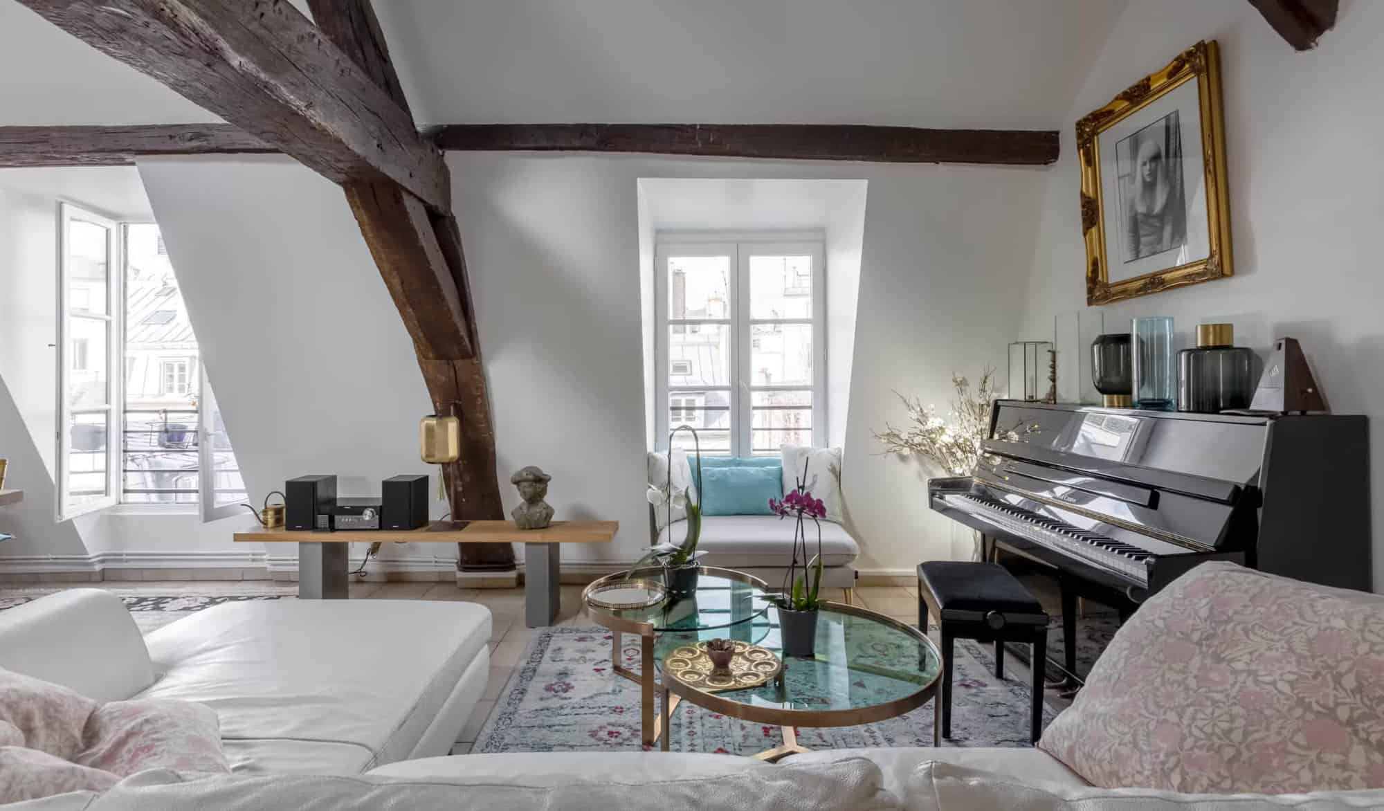 A Paris apartment with 2 windows overlooking the city. In it is a black piano and a white couch.