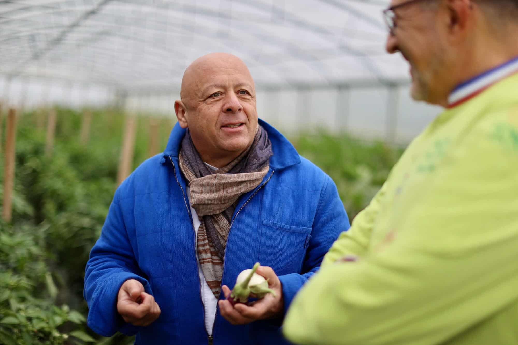 Chef Thierry Marx dressed in a blue winter jacket and beige scarf visits a greenhouse growing produce in Yvelines. 